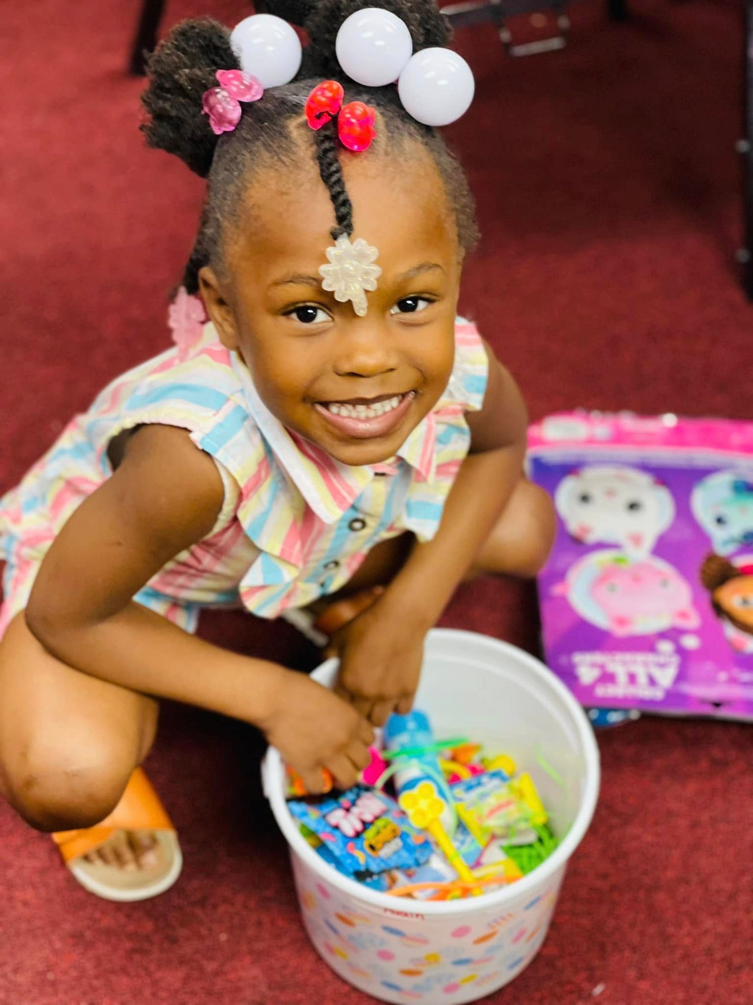 A smiling child crouches on a red carpet, holding a white bucket filled with colorful small toys and snacks.