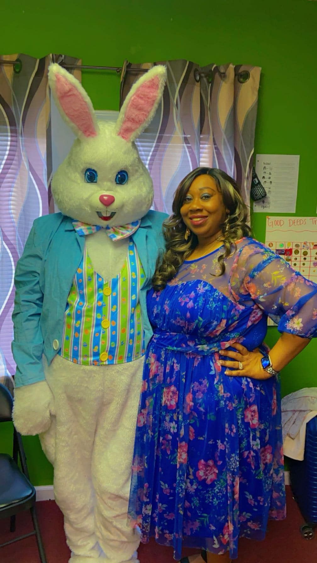 A person in a blue floral dress posing with a person in an Easter bunny costume against a green wall with curtains.