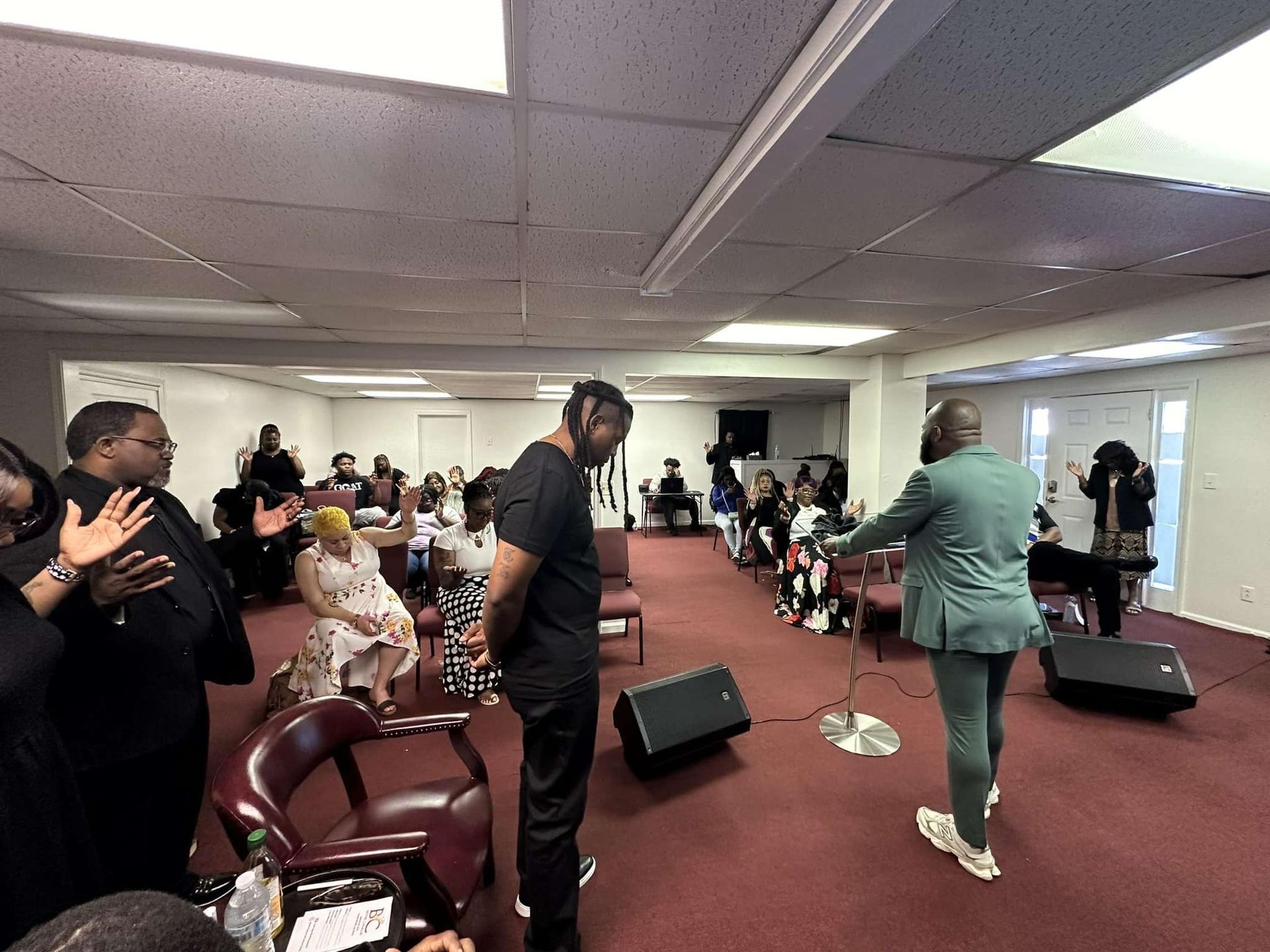 A group of people standing with hands raised in a room with a red carpet and ceiling lights, facing a speaker.