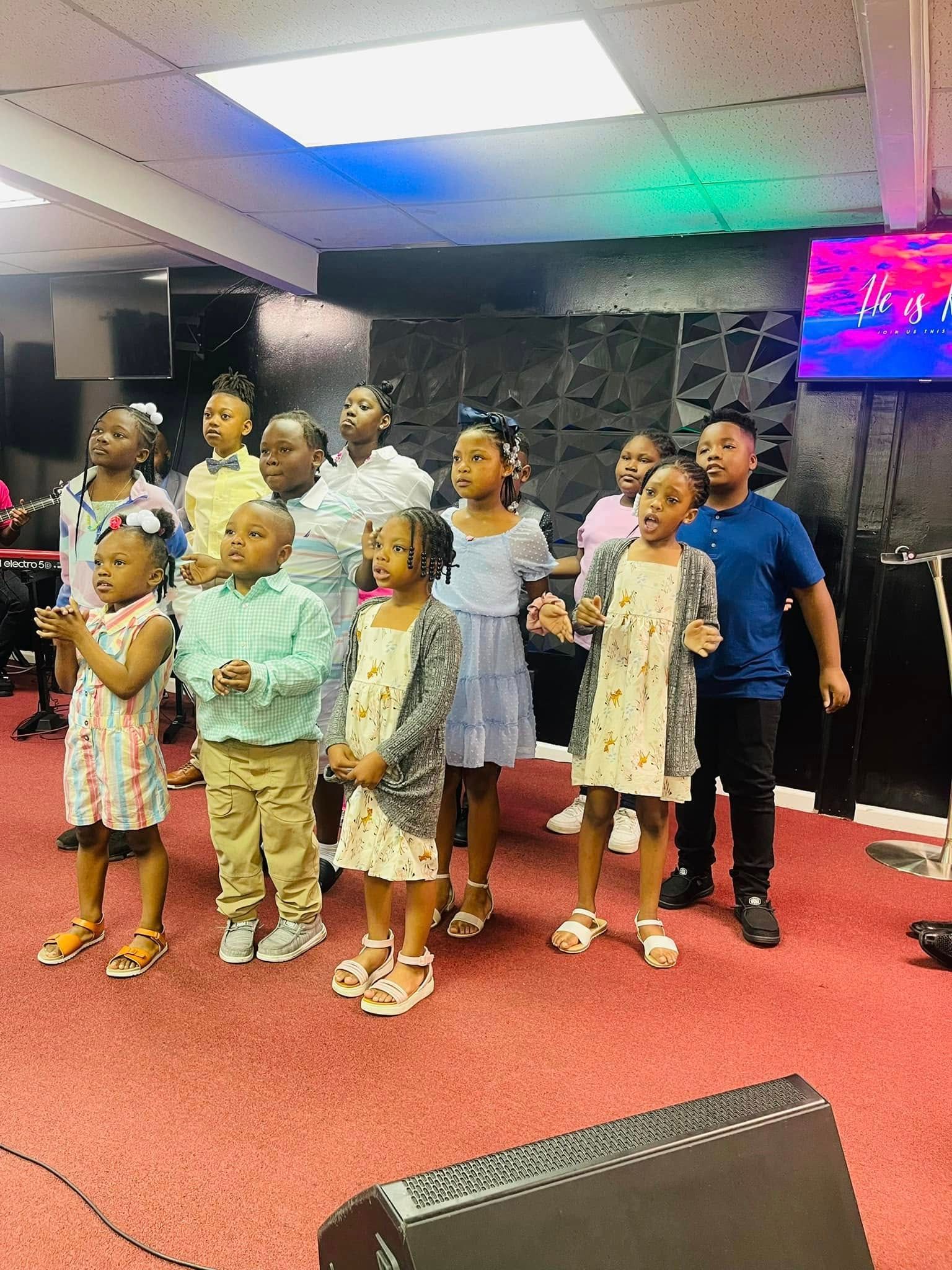 A group of children stand on a stage with red carpet, looking forward during a performance under bright indoor lights.