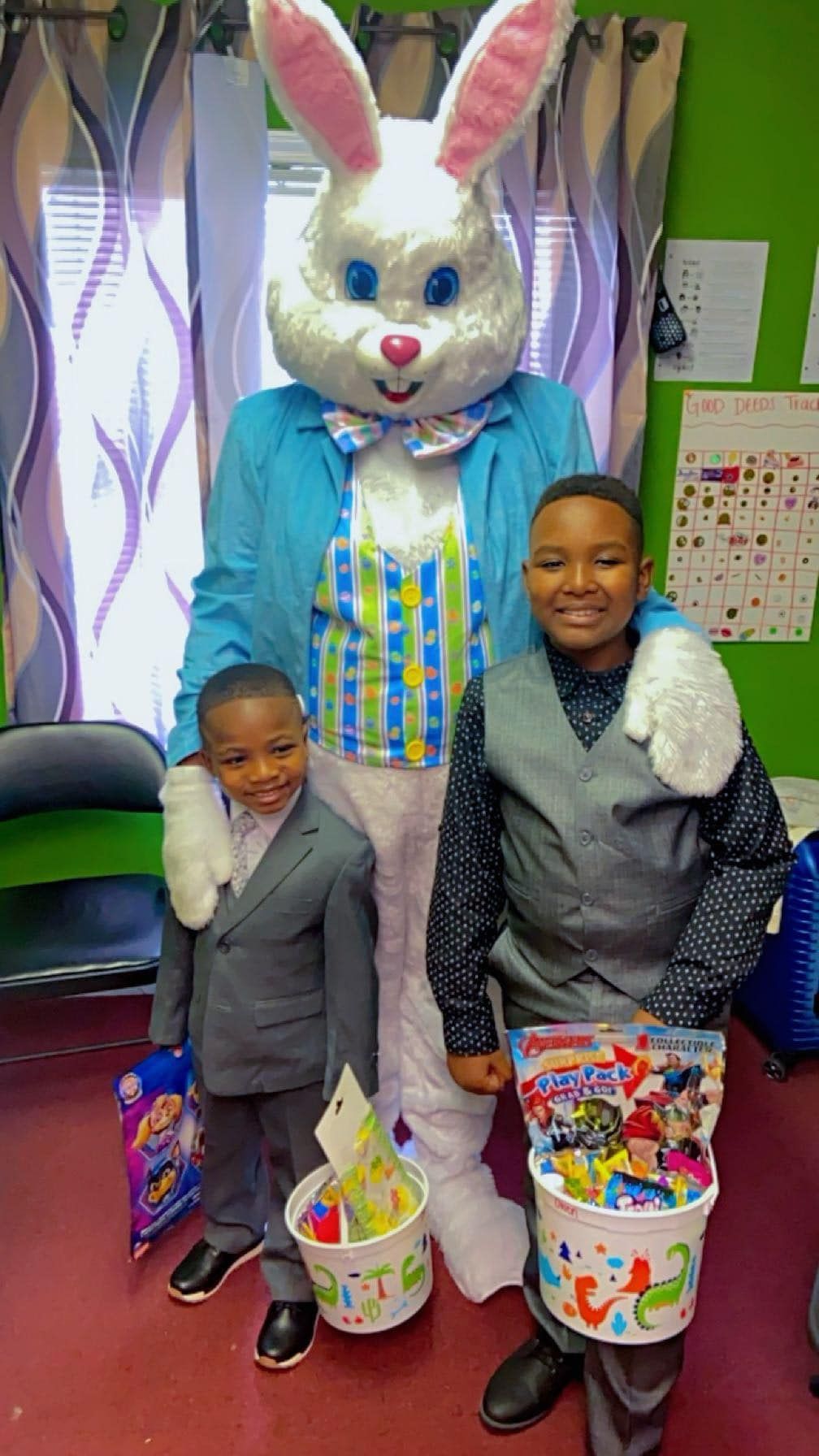 Two children in formal wear holding Easter baskets stand with an Easter Bunny mascot in an indoor setting.