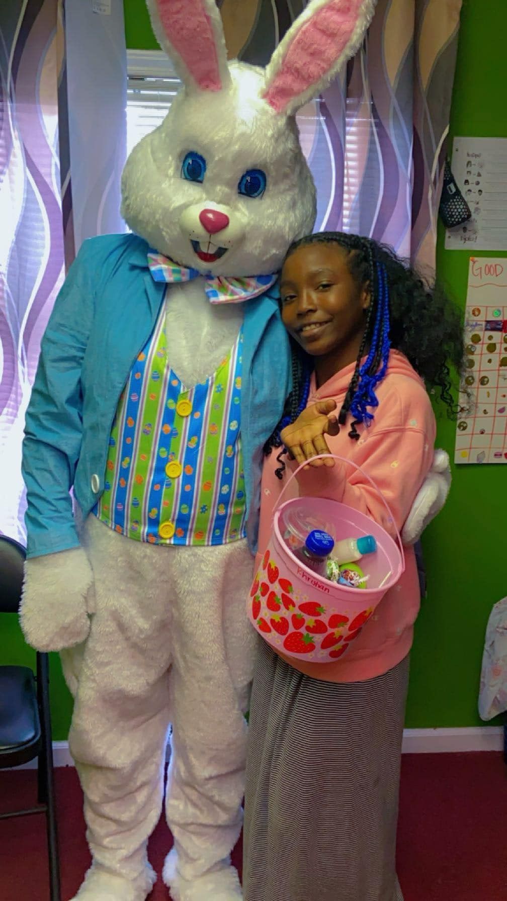 A person holding an Easter basket poses for a photo with a costumed Easter Bunny in a room with green walls.