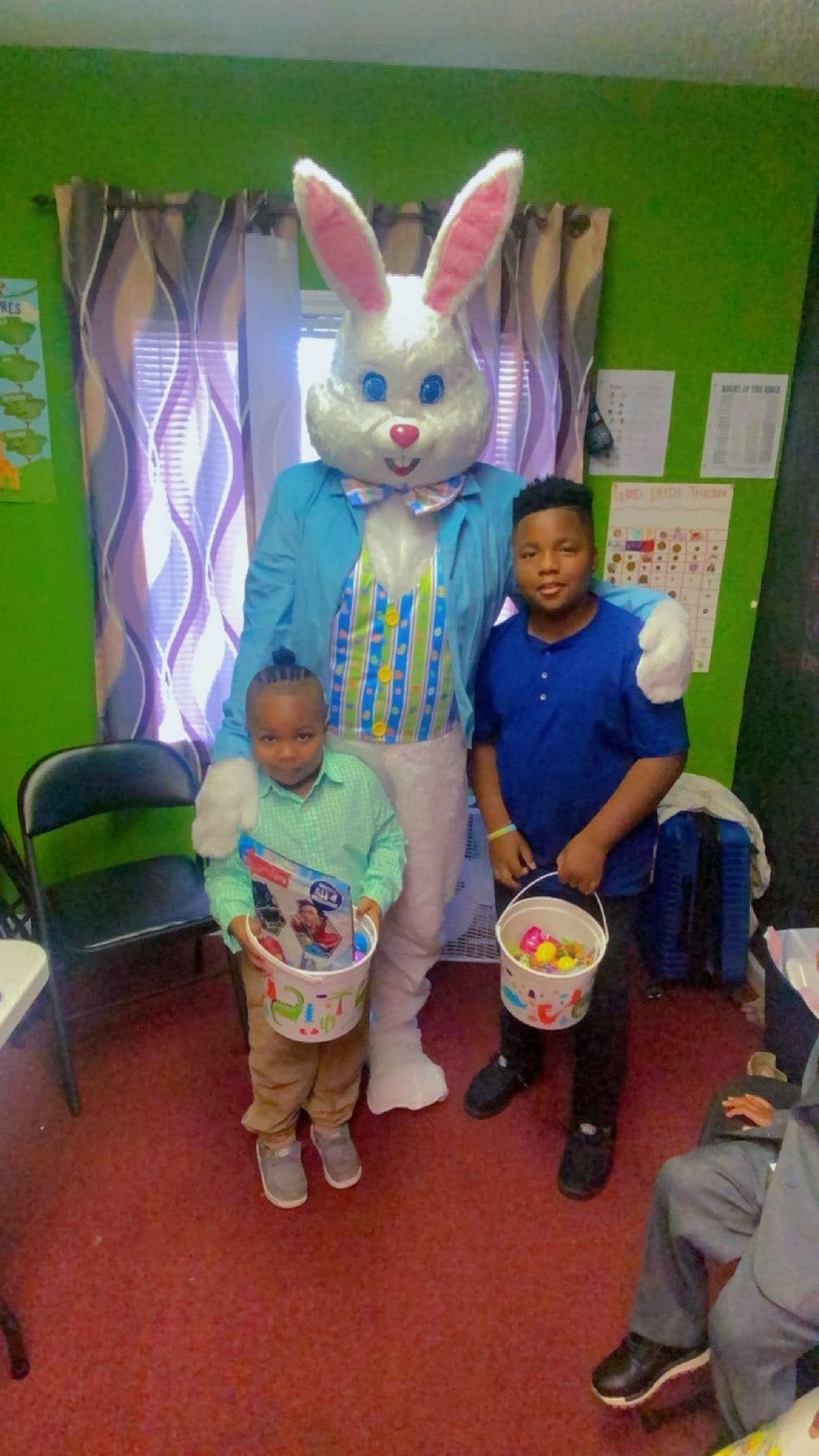 Two children hold Easter baskets while posing with a costumed Easter Bunny in a room with green walls and patterned curtains.