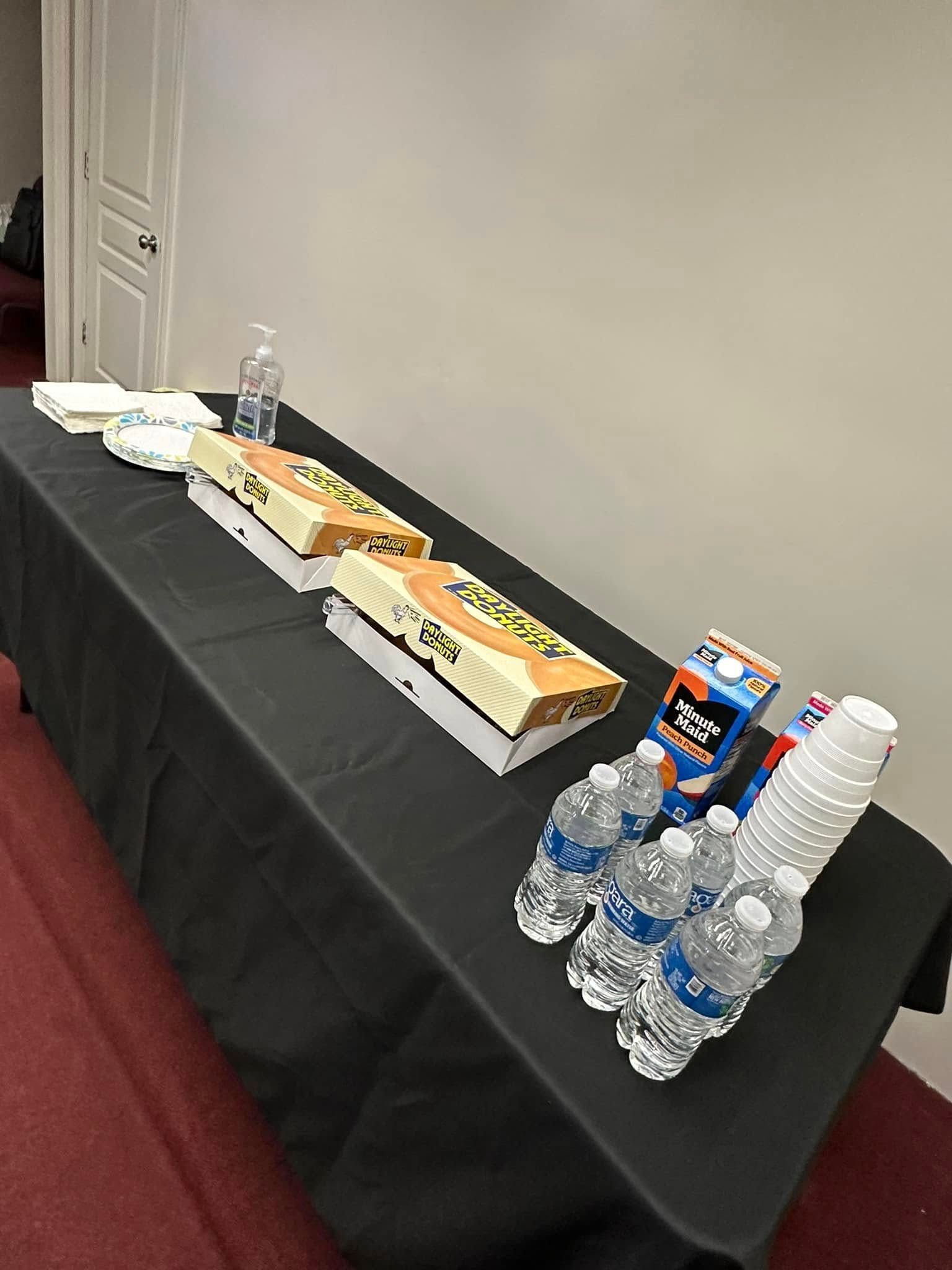 A black tablecloth-covered table with two pizza boxes, water bottles, cups, hand sanitizer, and plates.