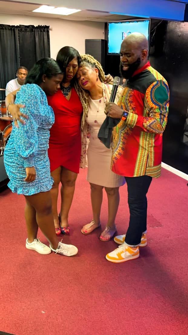 Four people in a circle holding hands and praying in a room with a red carpet floor.