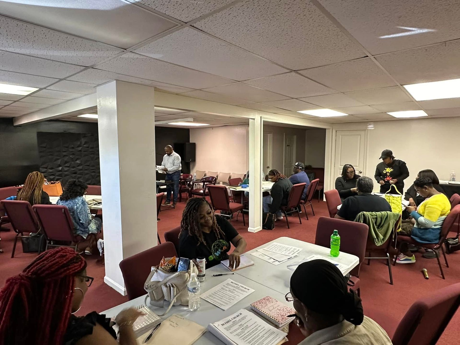 People sitting at tables in a community room for a meeting, with papers and supplies visible on the tabletops.