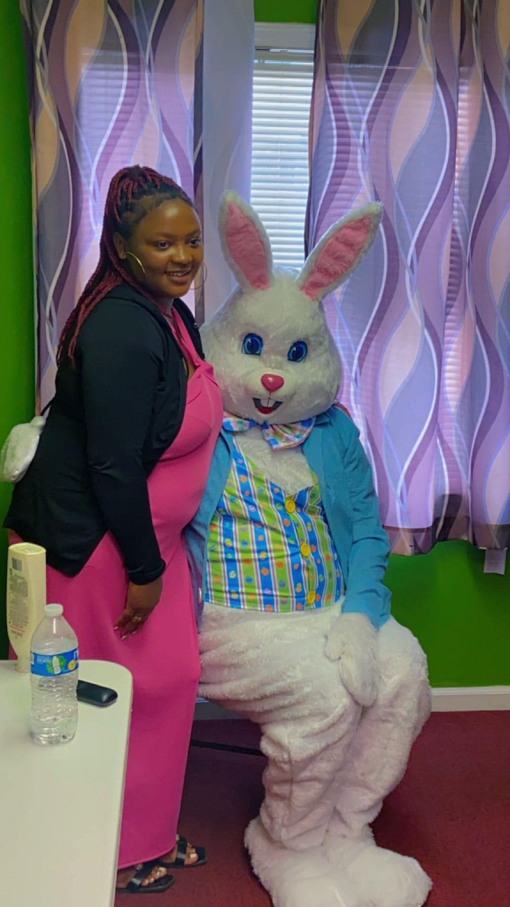 A smiling person in a pink dress and black cardigan stands next to a person in a large, white Easter bunny costume.