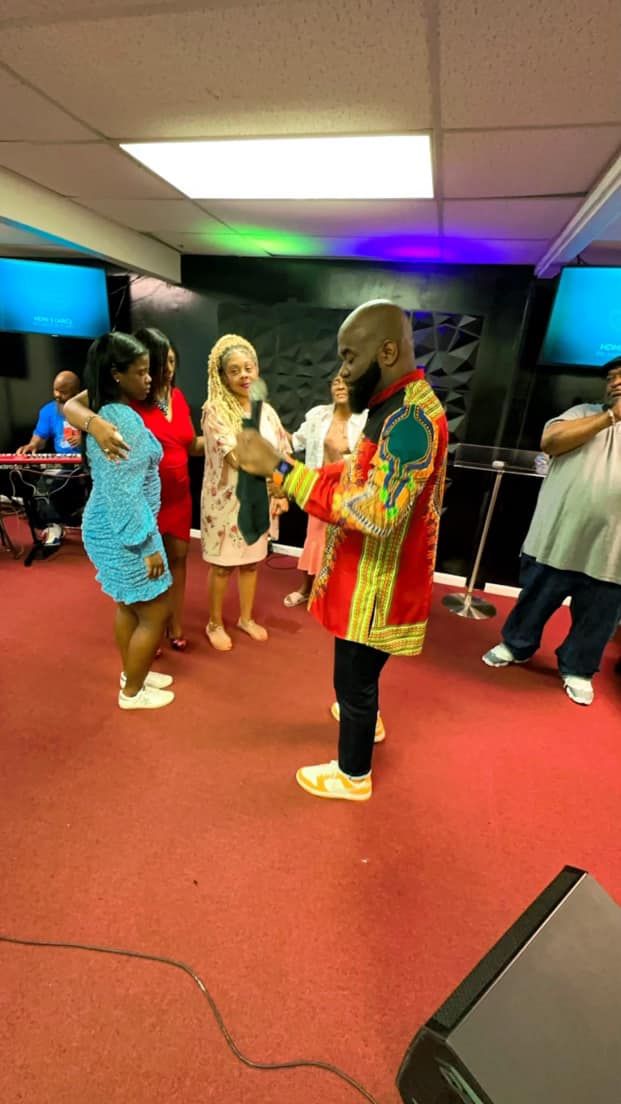 A person in a patterned red and gold jacket speaks into a microphone while others stand nearby in a room with red carpet.