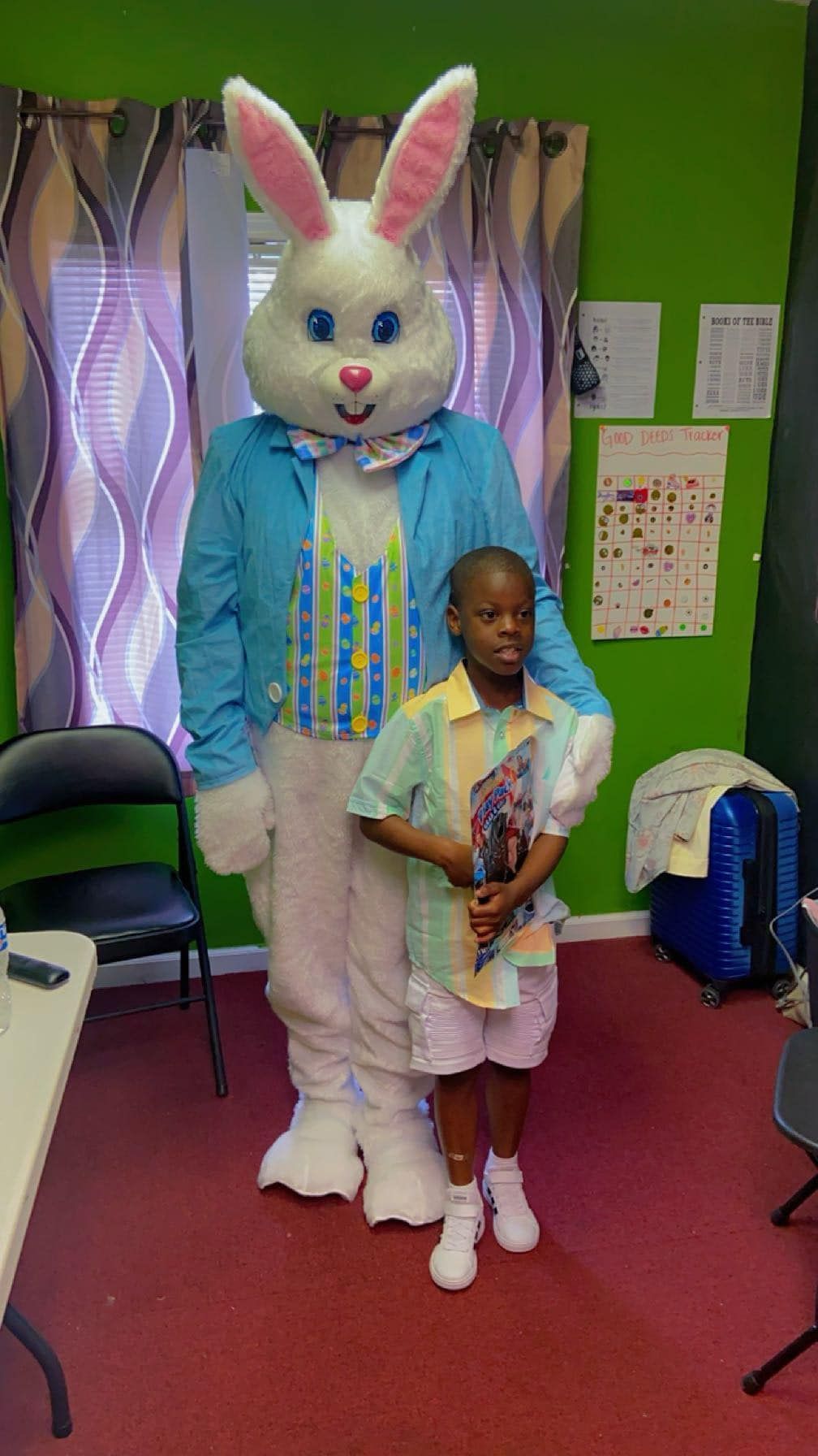 A child stands next to a person in a full-body Easter Bunny costume wearing a blue blazer and vest in an indoor room.
