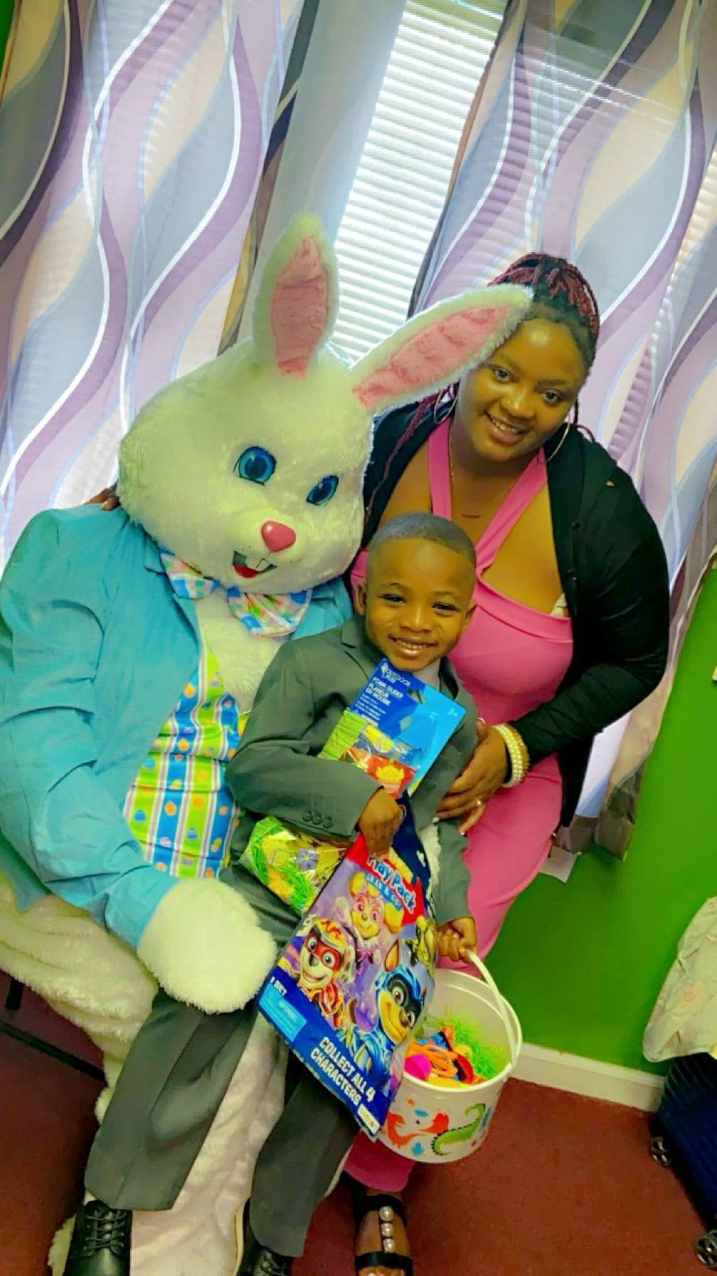 A smiling child holds a toy and basket while posing with an Easter Bunny mascot and an adult in a pink top.