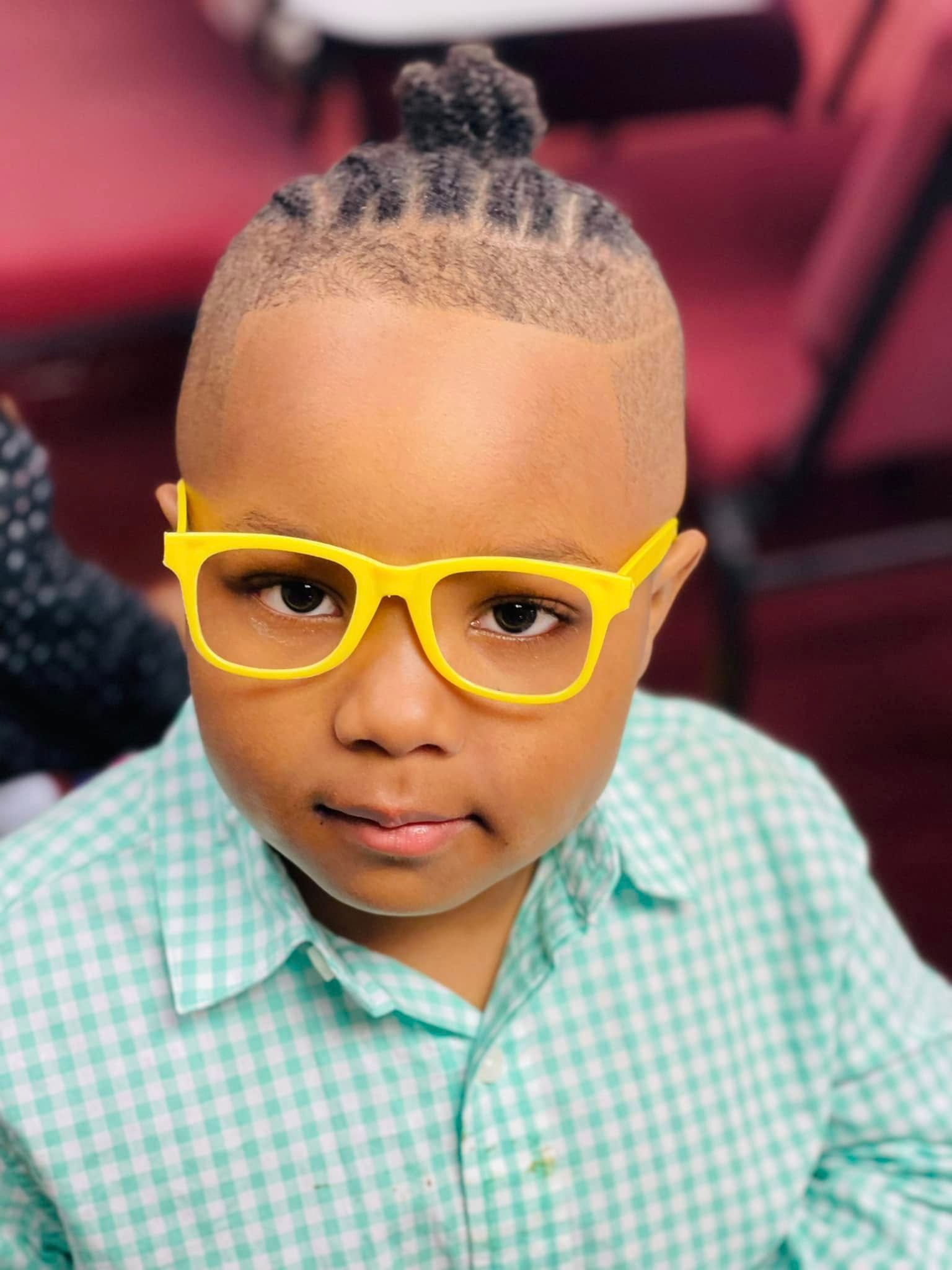 A person with a braided top-knot hairstyle and faded sides, wearing bright yellow glasses and a green checkered shirt.