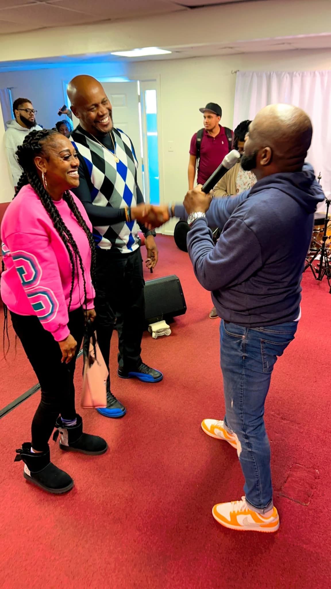 A man with a microphone interviews a woman and a man in a sweater at an indoor event with a red carpet.