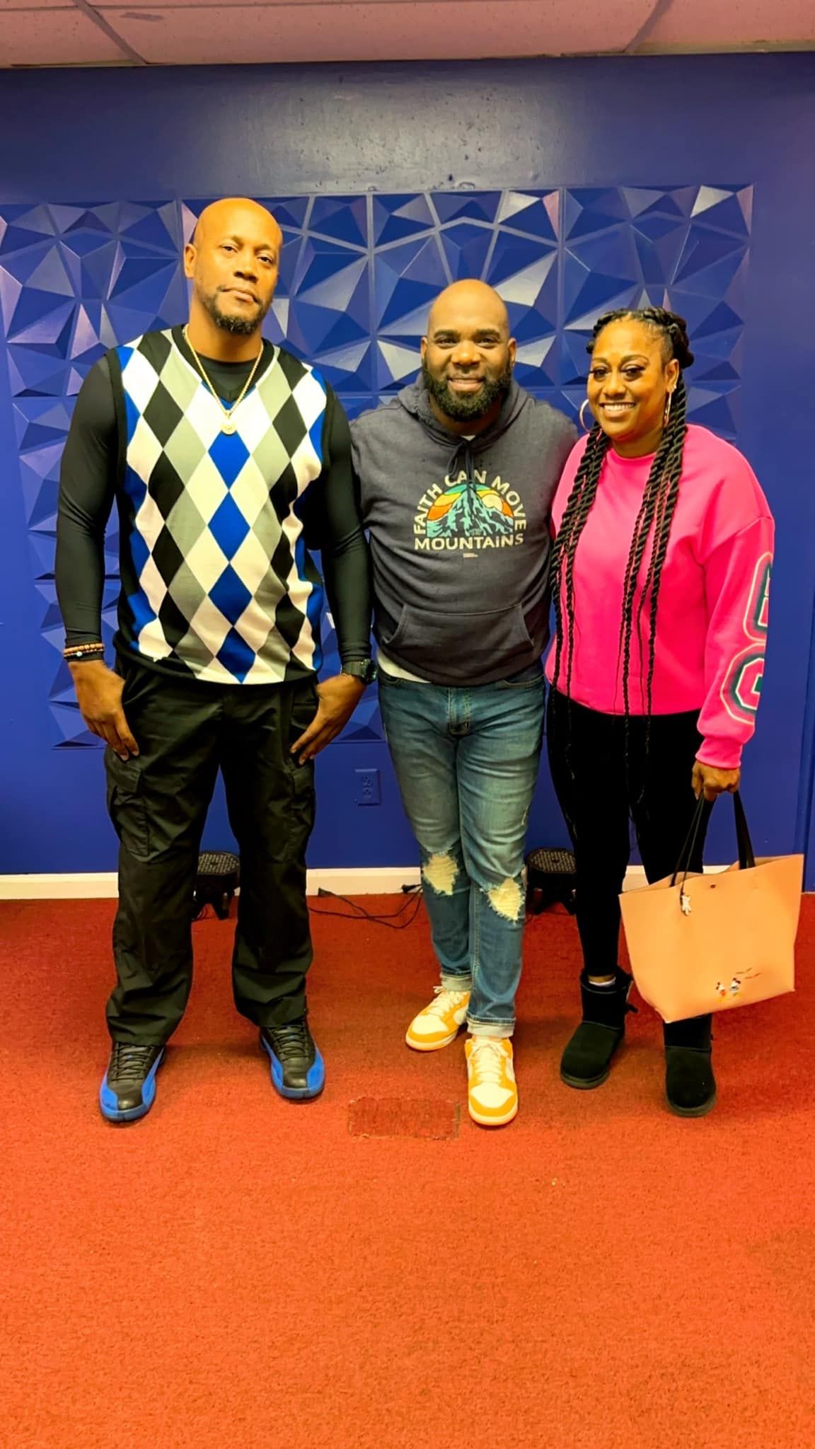 Three people stand together smiling against a textured blue wall with a red carpet floor.