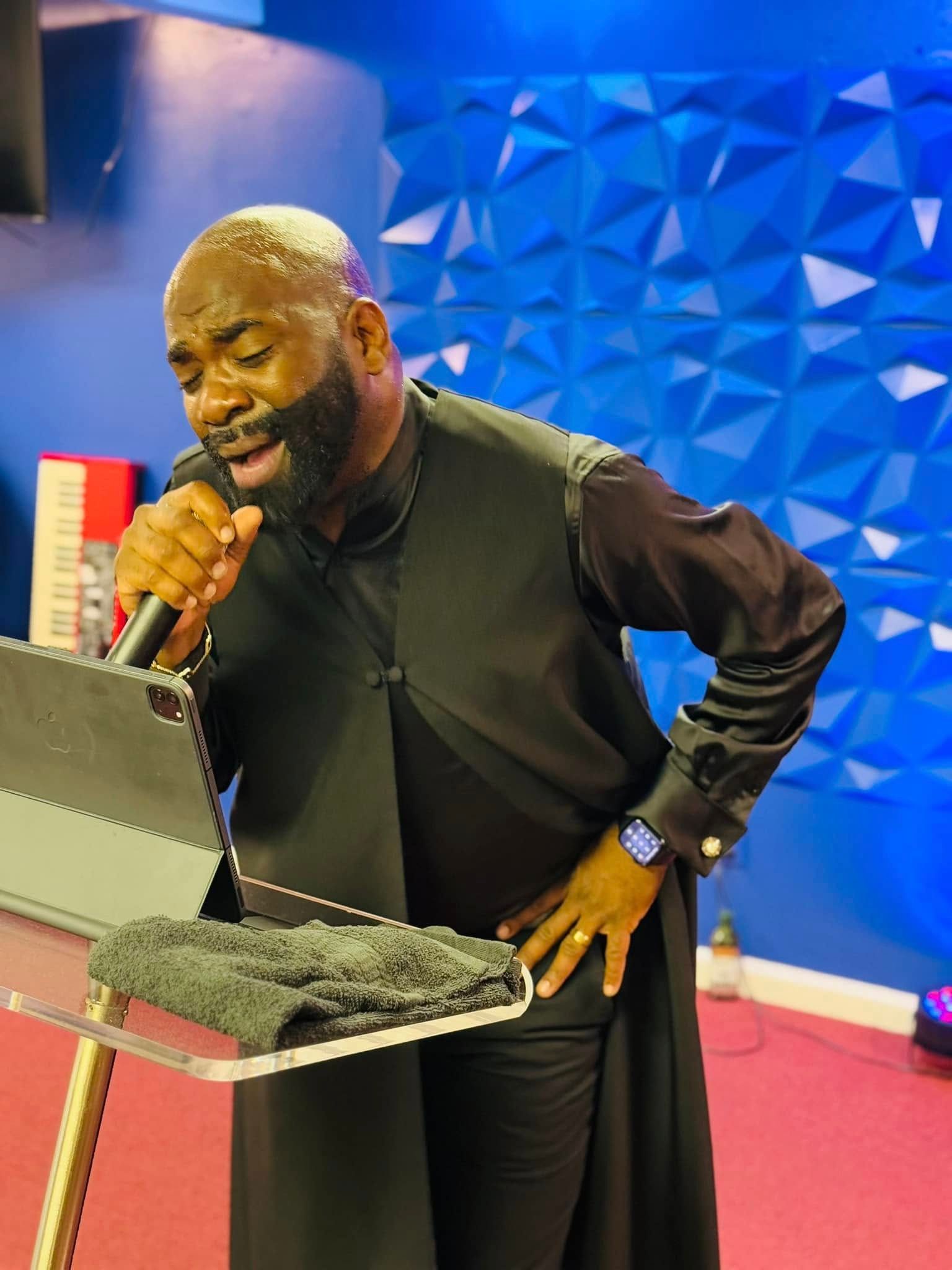 A person singing into a microphone while looking at a tablet on a stand, standing before a blue geometric wall.