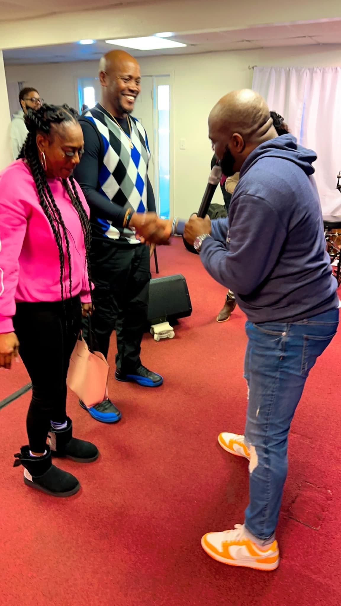 A person in a blue hoodie talks into a microphone while shaking hands with another person in a sweater indoors.