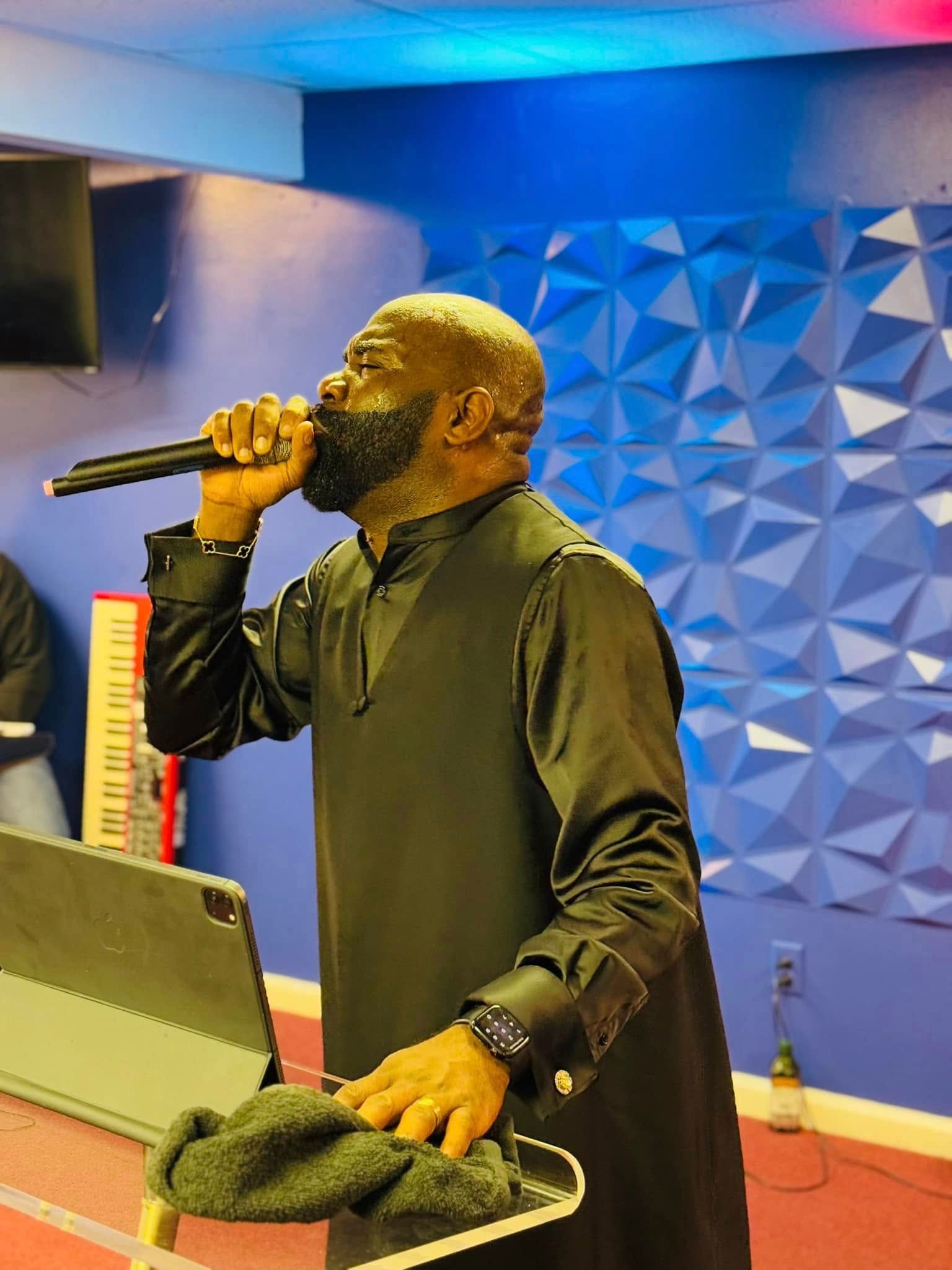 A person in a black shirt holds a microphone to their mouth while standing behind a podium in a blue studio room.