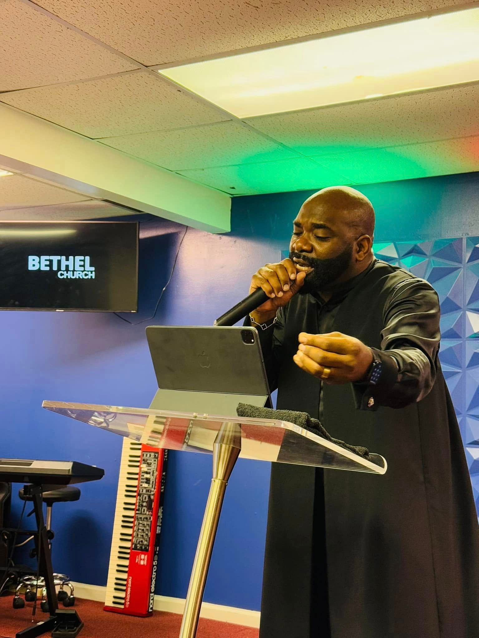 A speaker stands at a clear lectern with a tablet, holding a microphone and gesturing while at Bethel Church.