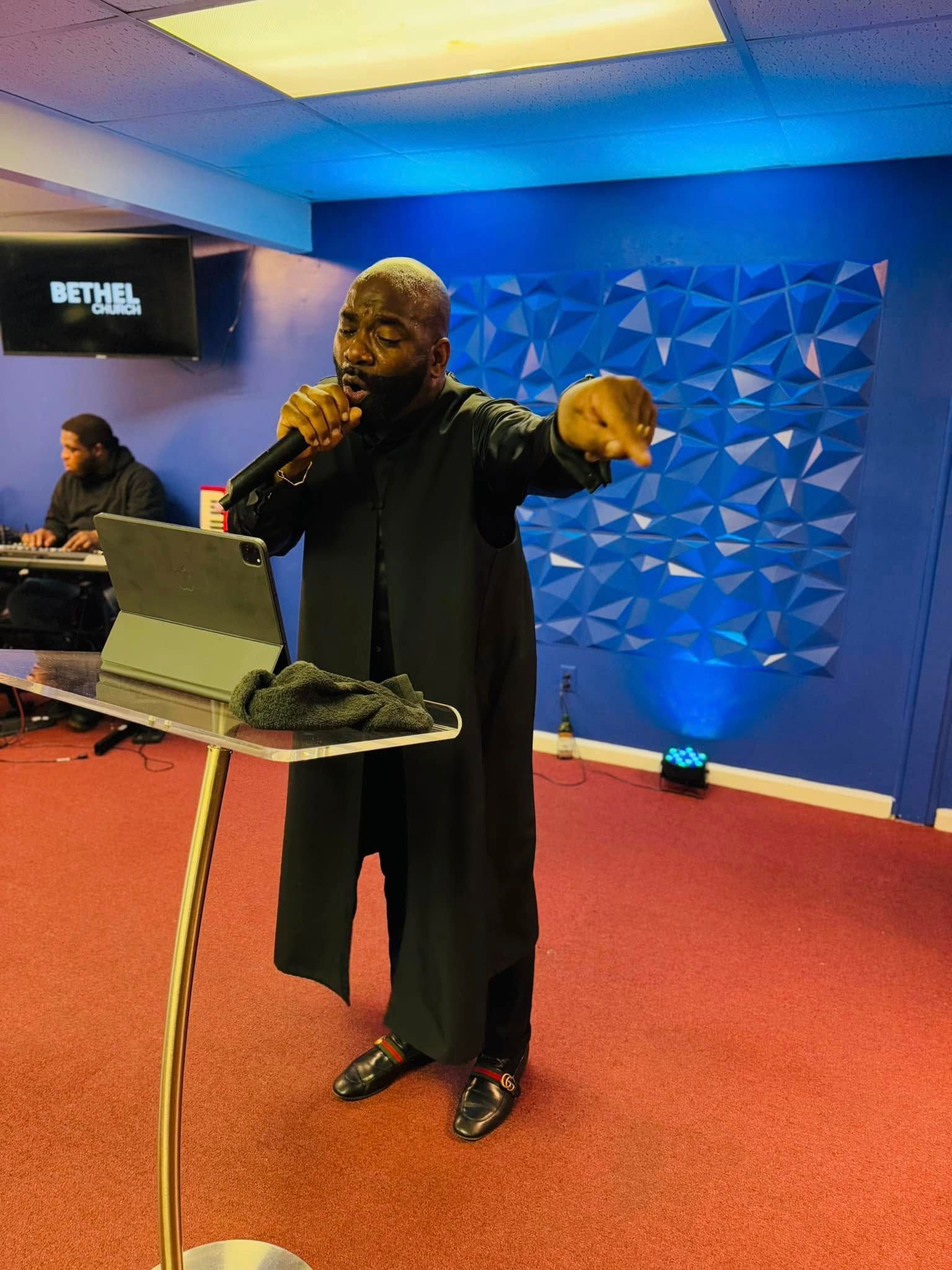 A person in a black coat stands at a podium holding a microphone and gesturing, with a blue wall behind them.
