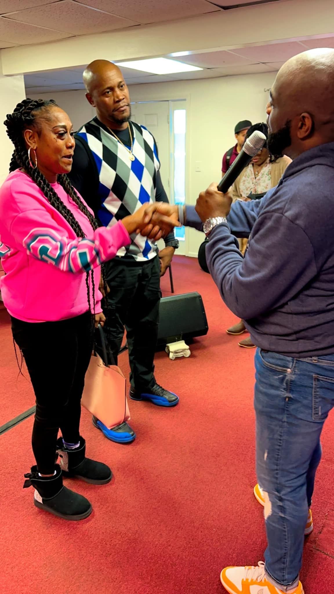 A person in a blue shirt holds a microphone to someone in a pink shirt who is shaking hands with someone in a sweater.