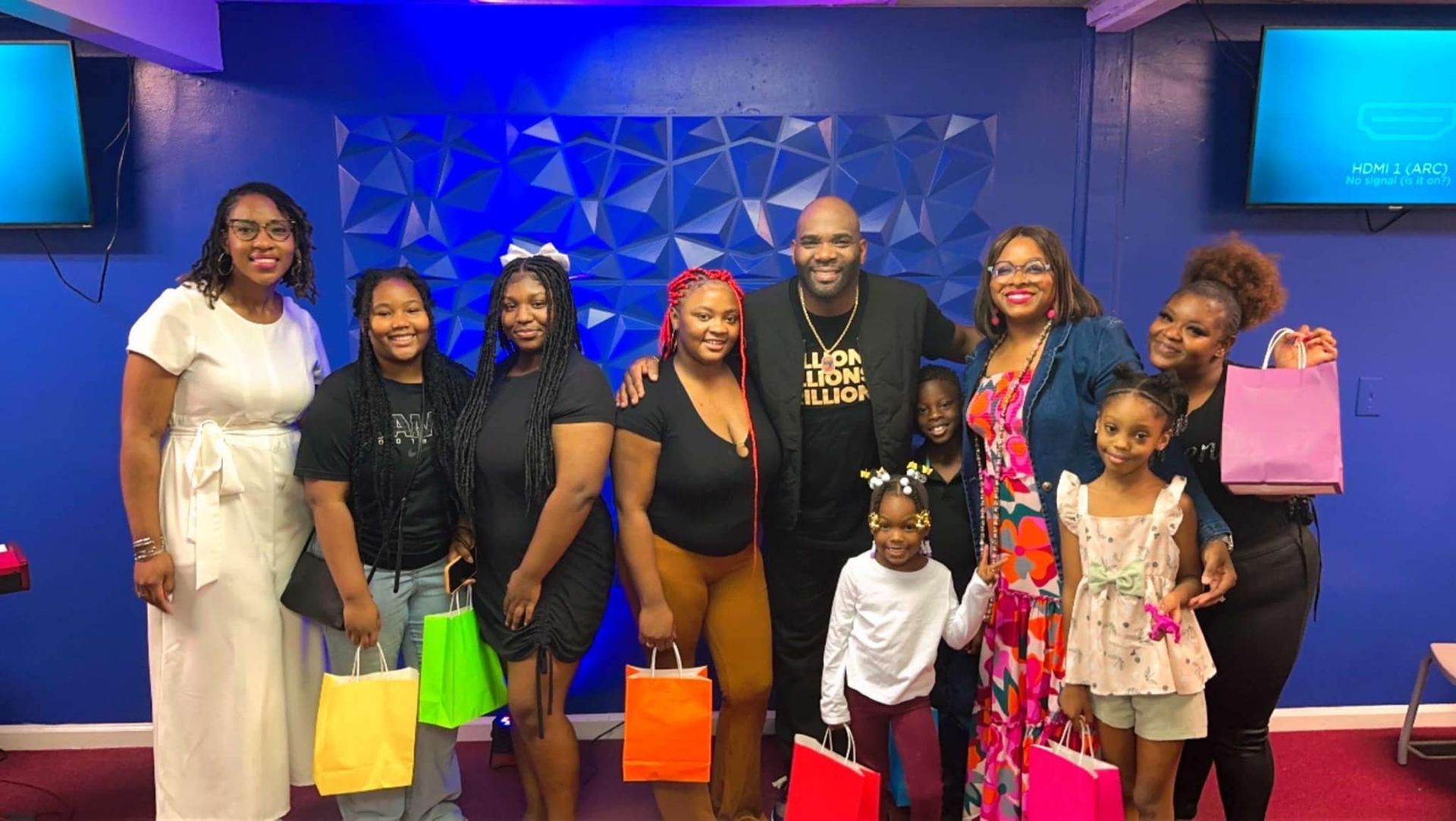 A group of people standing together in a room with blue walls, holding colorful gift bags and smiling at the camera.