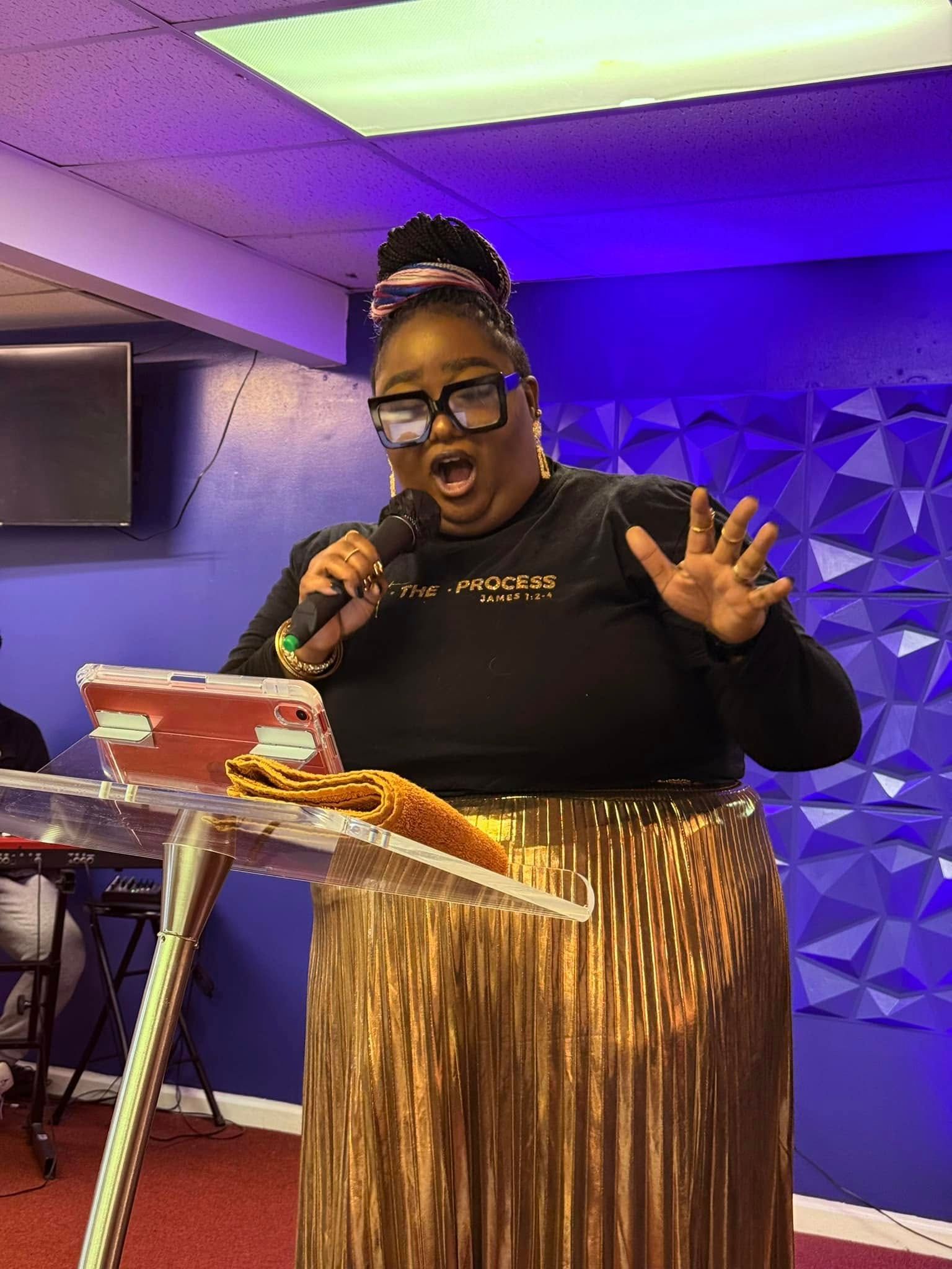 A person speaking into a microphone at a podium, wearing glasses and a metallic gold pleated skirt against a blue background.