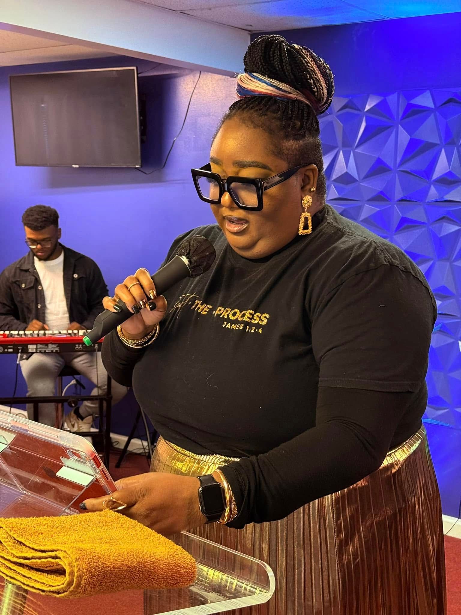 A person in a black shirt and gold skirt speaks into a microphone at a podium with a keyboard player in the background.