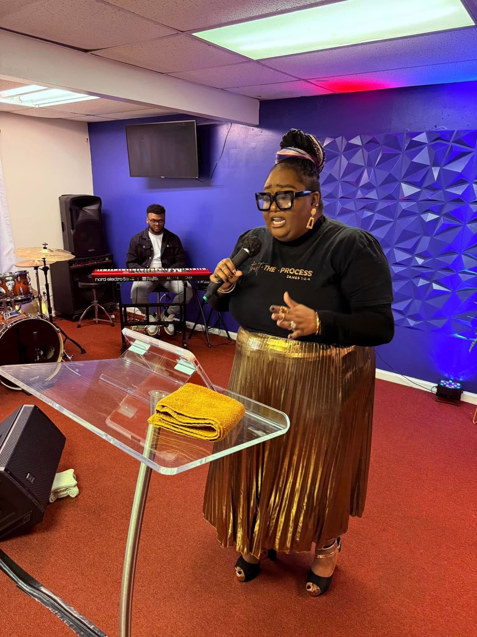 A person stands at a transparent podium speaking into a microphone, with a keyboardist in the background on a blue stage.