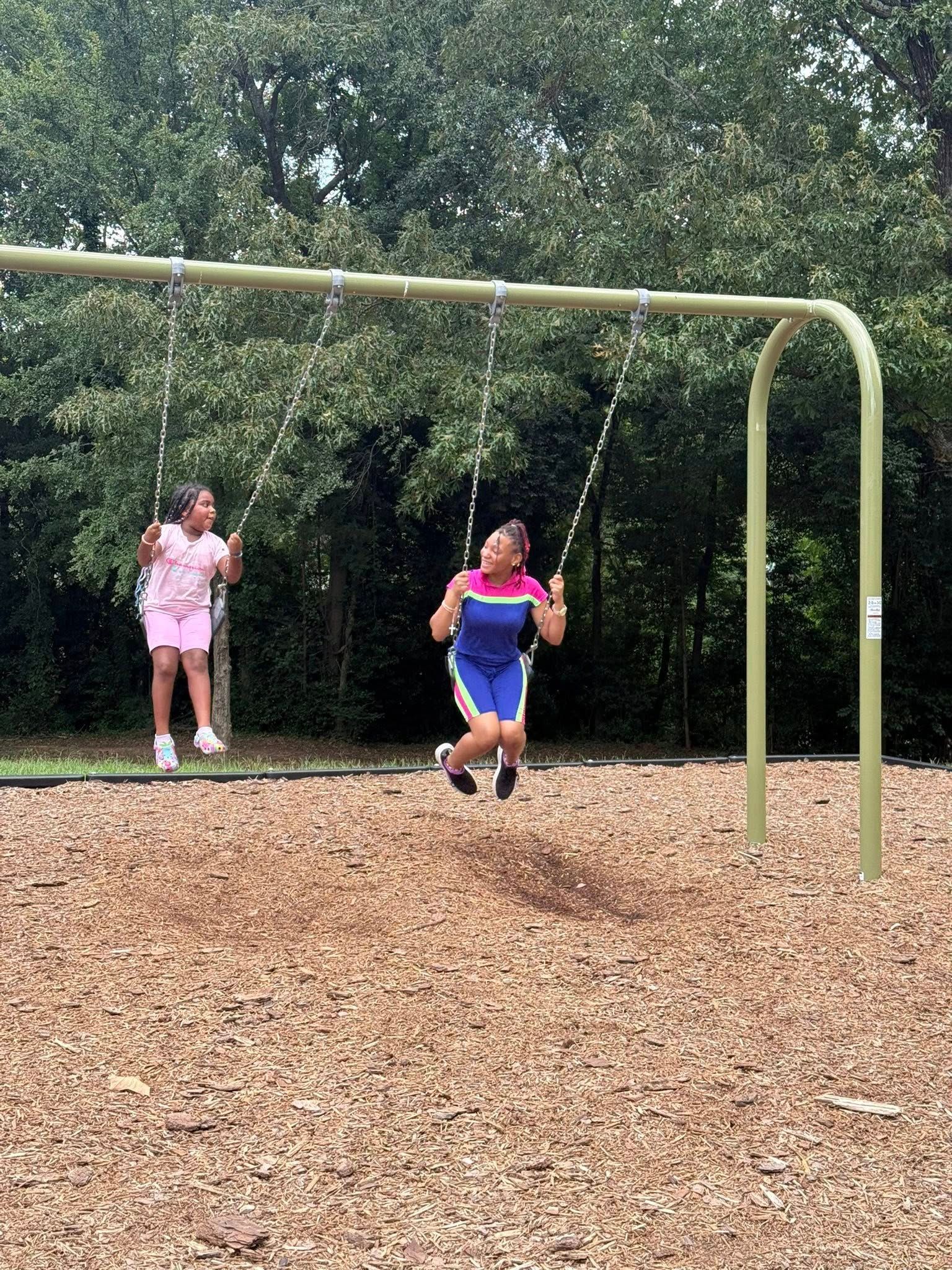 Two people swing on a playground set above wood chips in front of a line of trees.