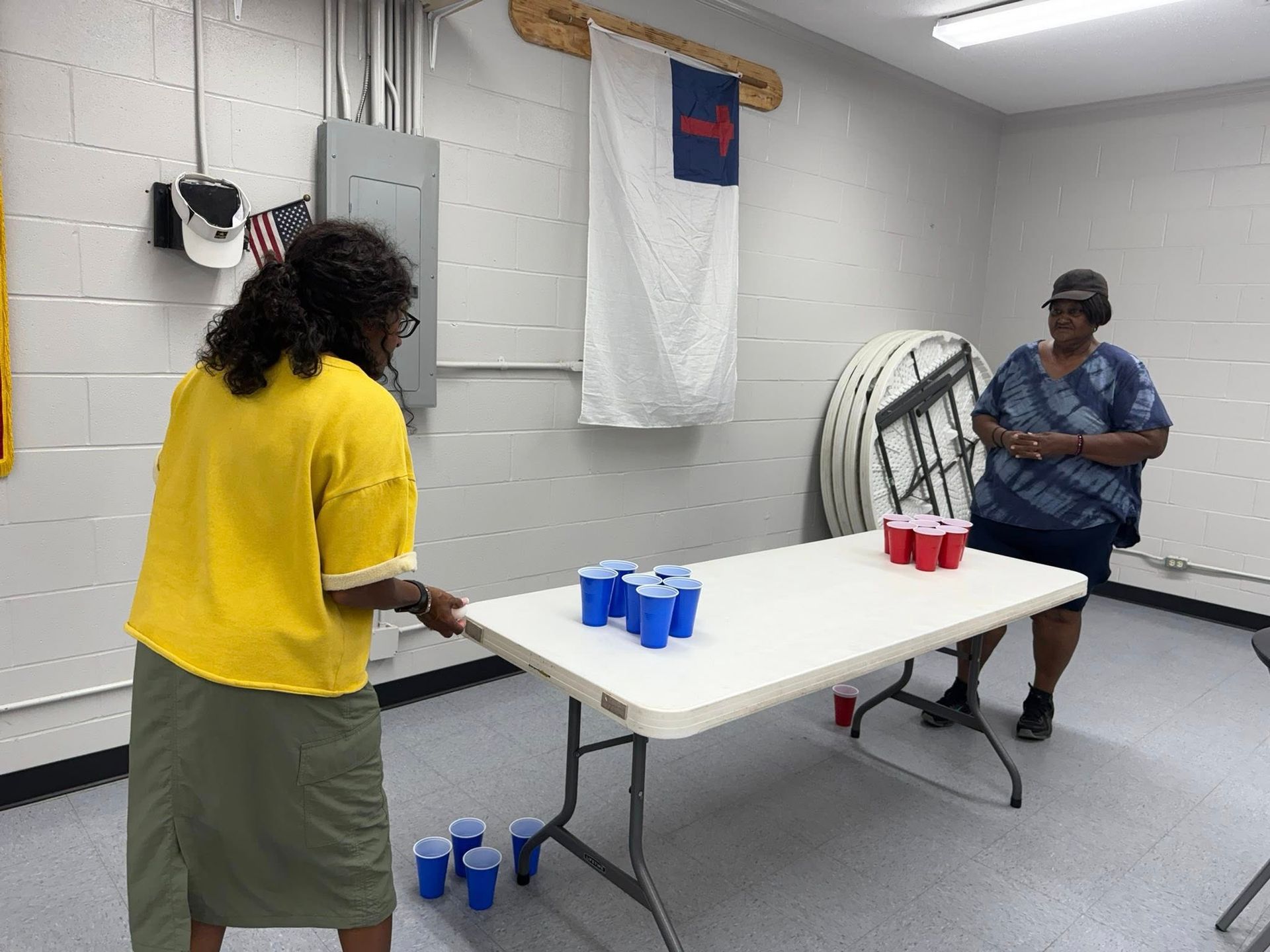 Two people stand at opposite ends of a folding table playing a cup-tossing game in an indoor room with cinder block walls.