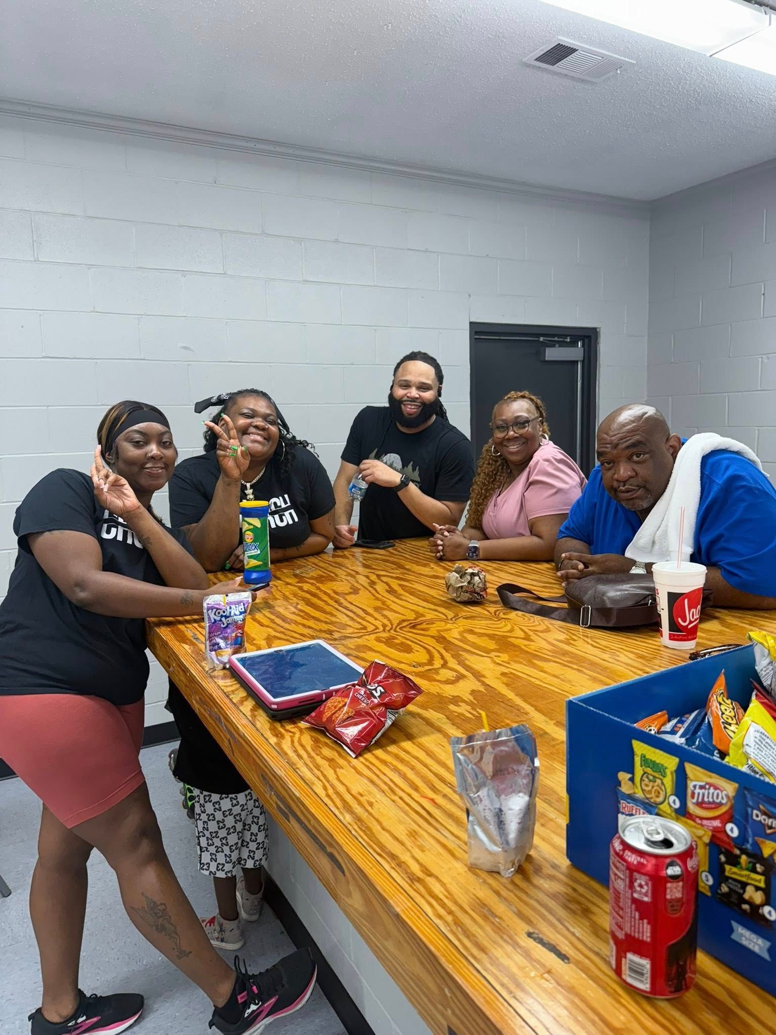 Five people sit around a long wooden table in a room with gray block walls, smiling at the camera with snacks and drinks.
