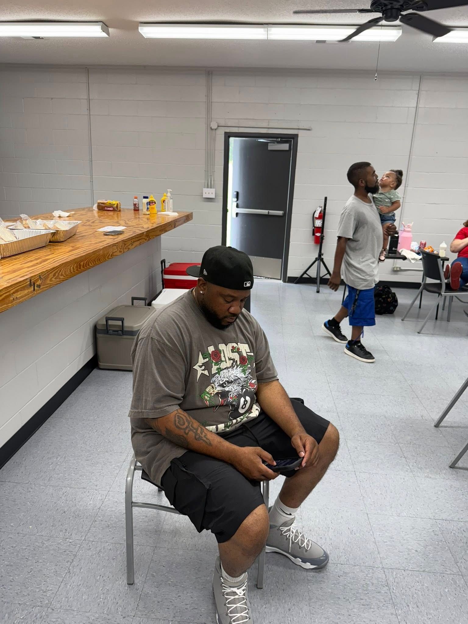 A person sits on a chair looking at a phone, while another person carries a child in the background of a room.