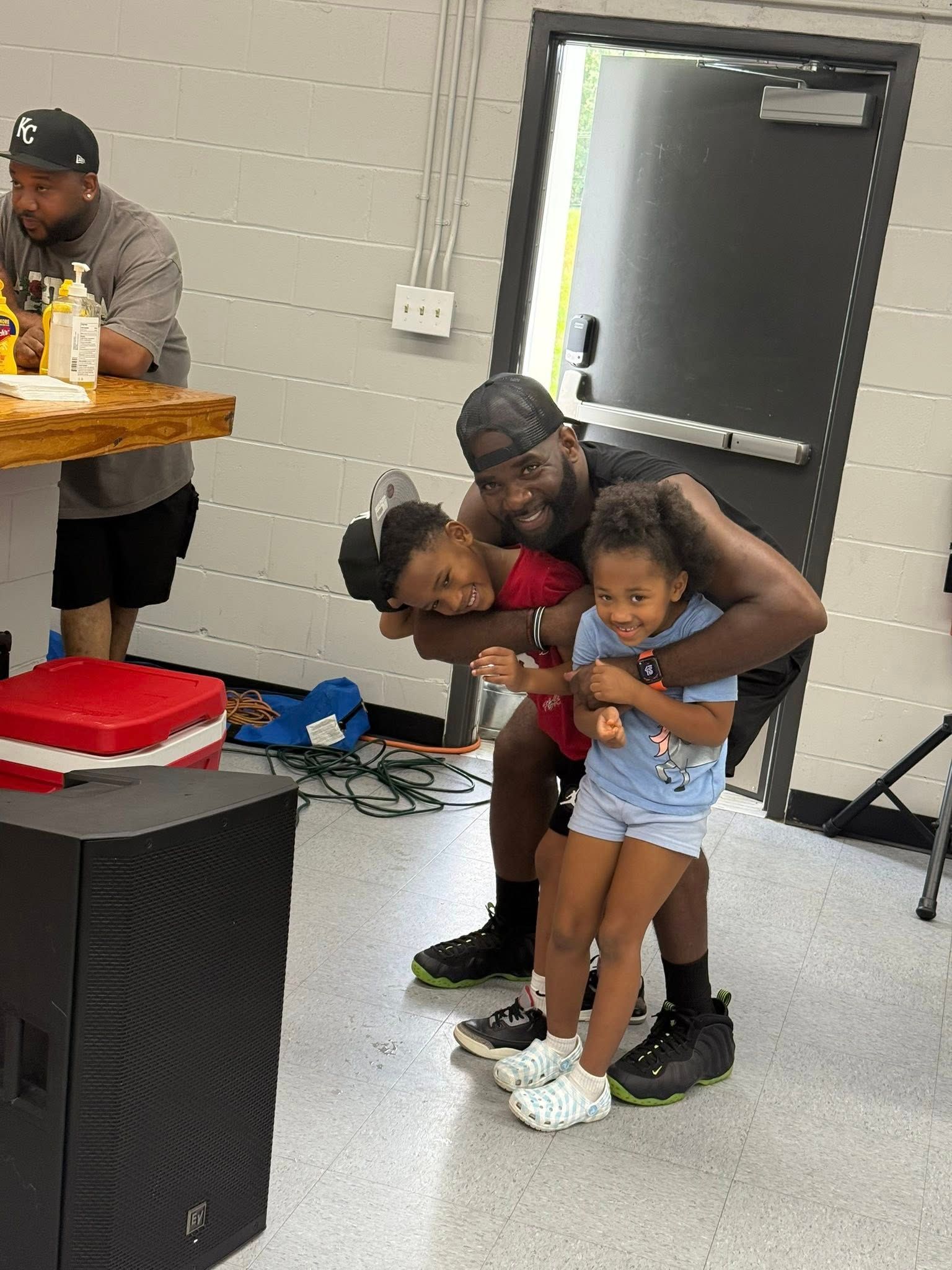 A man crouches in an indoor room, hugging two children while smiling, as another person sits at a table nearby.