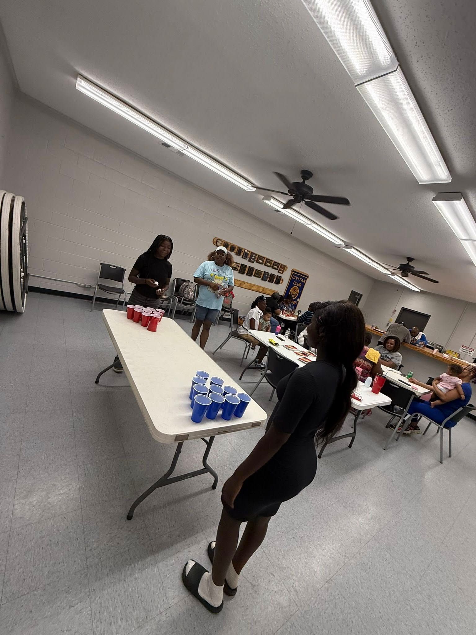 People play a game with blue plastic cups arranged on a folding table in a brightly lit, spacious community room.