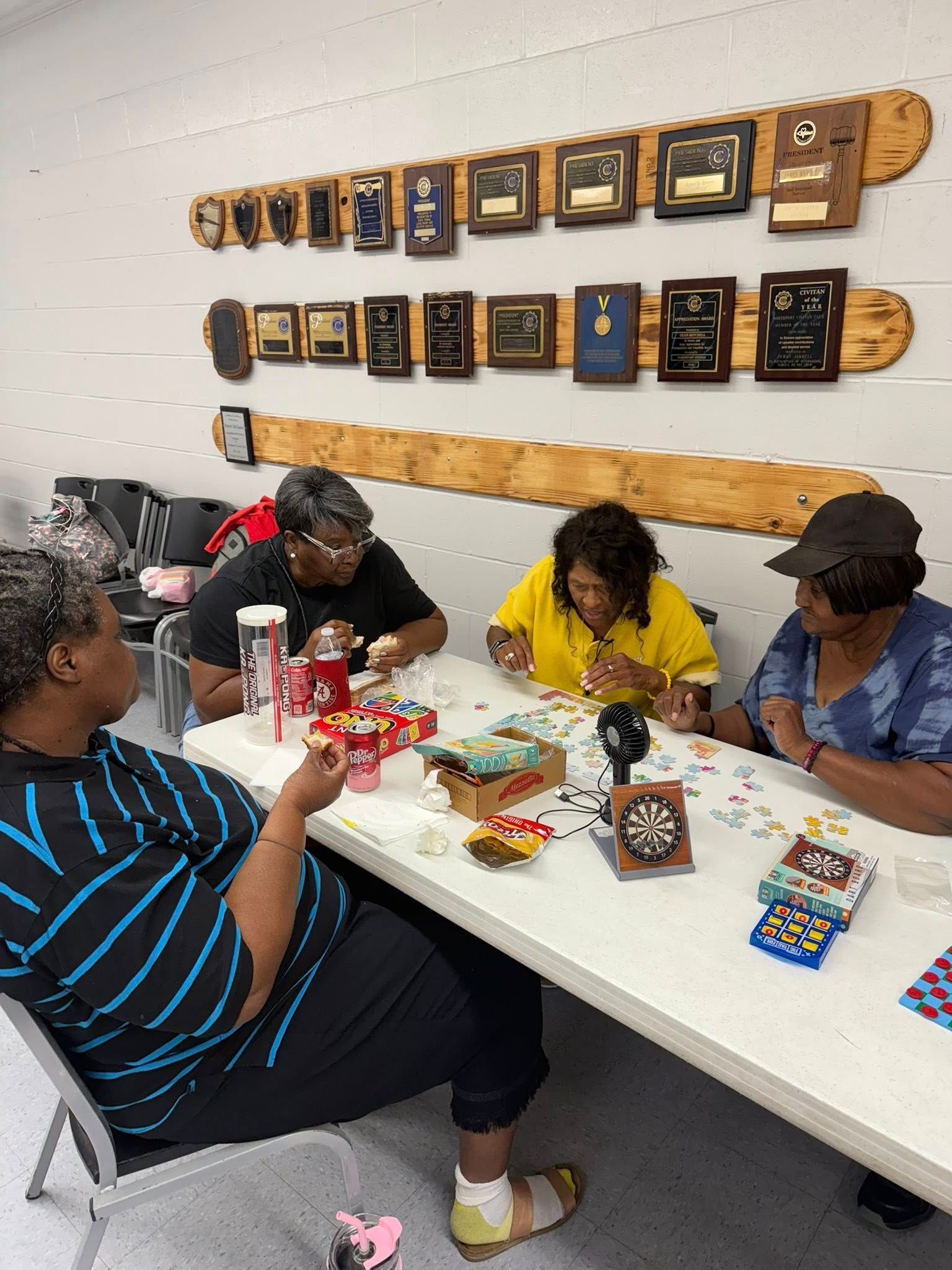 Four people sit around a table working on a jigsaw puzzle in a room with numerous award plaques on the wall.