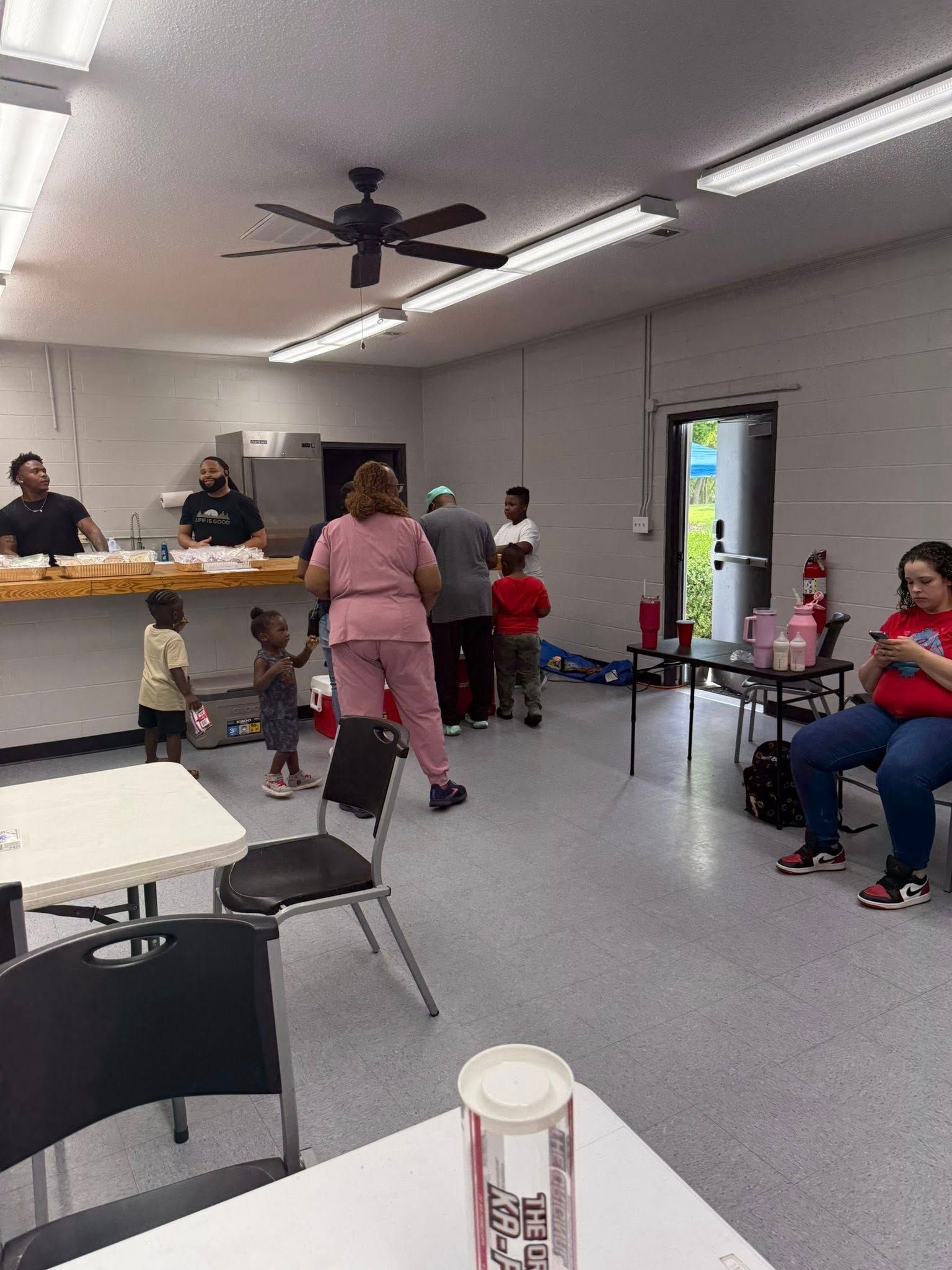 People gather in a bright community room with tables, a counter, and a doorway leading outside.