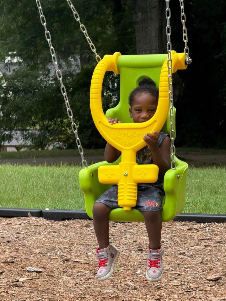 A child sits in a lime-green, toddler-safe swing with a yellow T-bar at a park with wood chips on the ground.