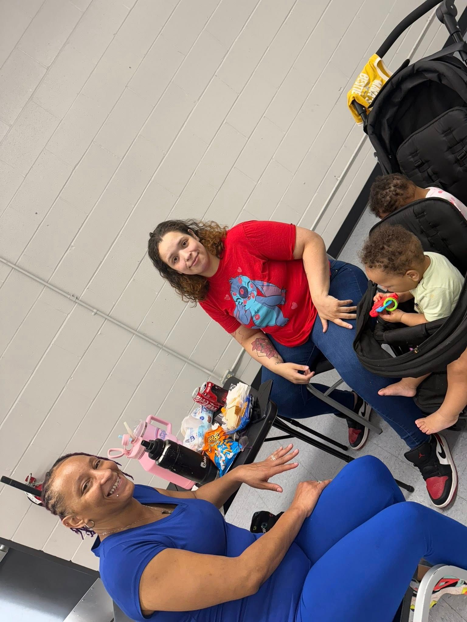 Two people sit near a stroller holding a child, with snacks on a table between them, in an indoor setting.