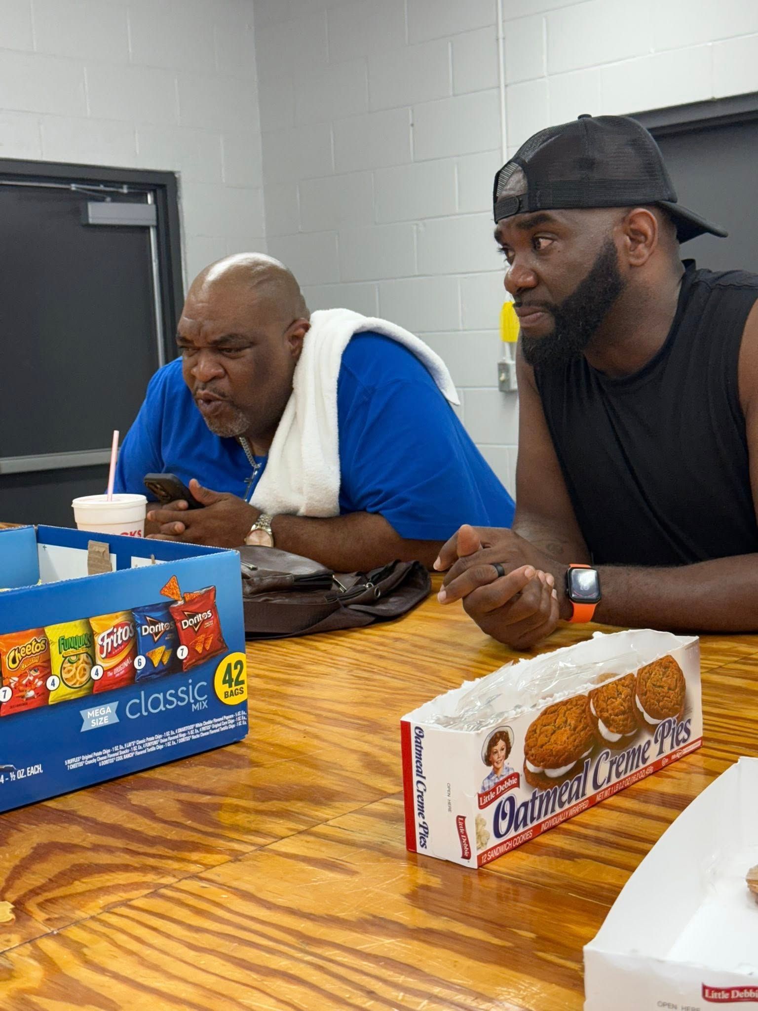 Two people sitting at a table with snack boxes and drinks, appearing to be in an indoor break room or office setting.
