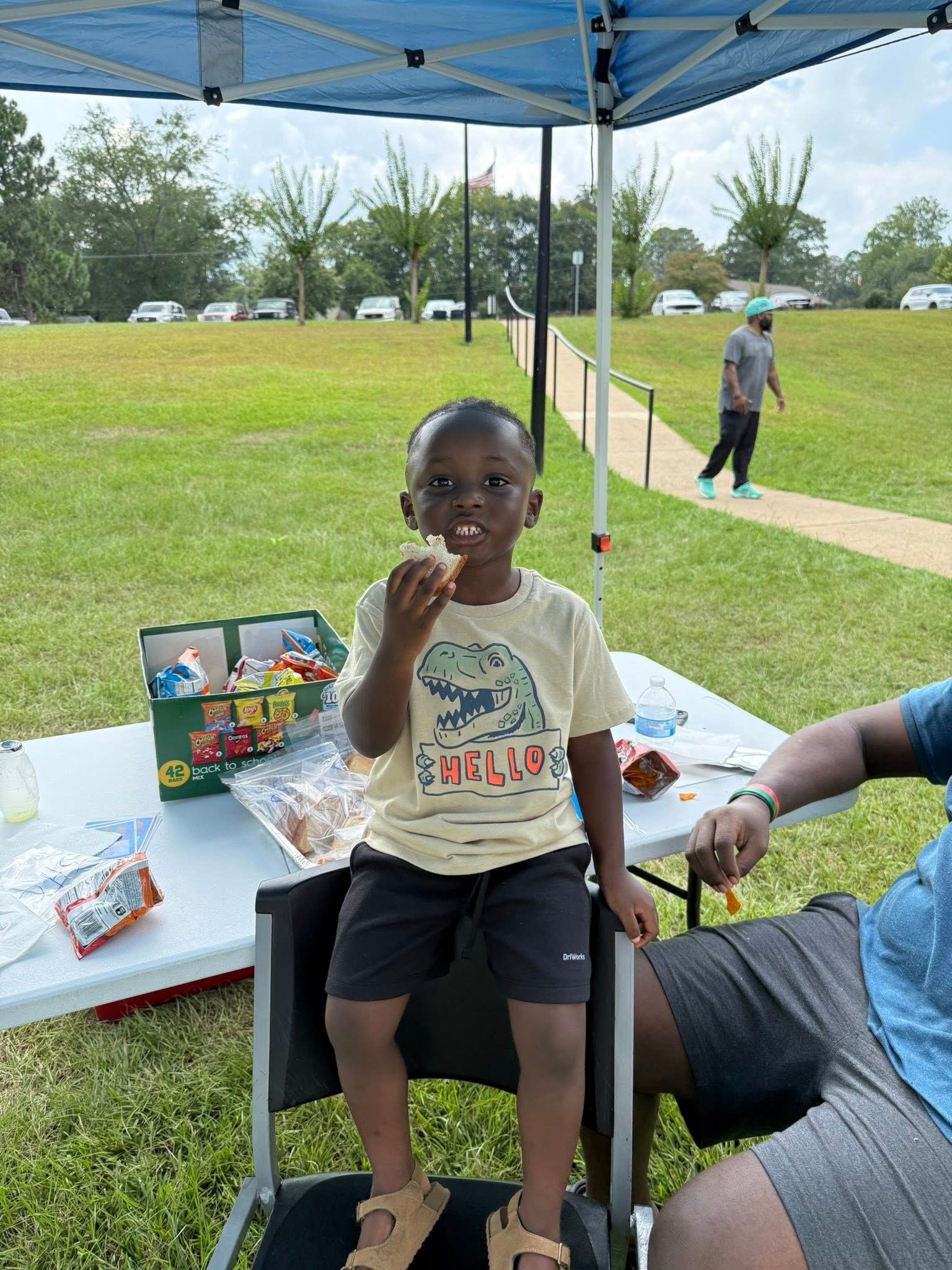 A child in a dinosaur t-shirt sits on a chair eating under a blue canopy at an outdoor park event.