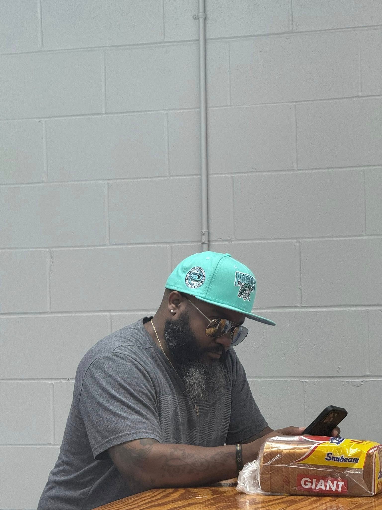 A bearded individual wearing a teal baseball cap sits at a wooden table looking at a smartphone next to a loaf of bread.