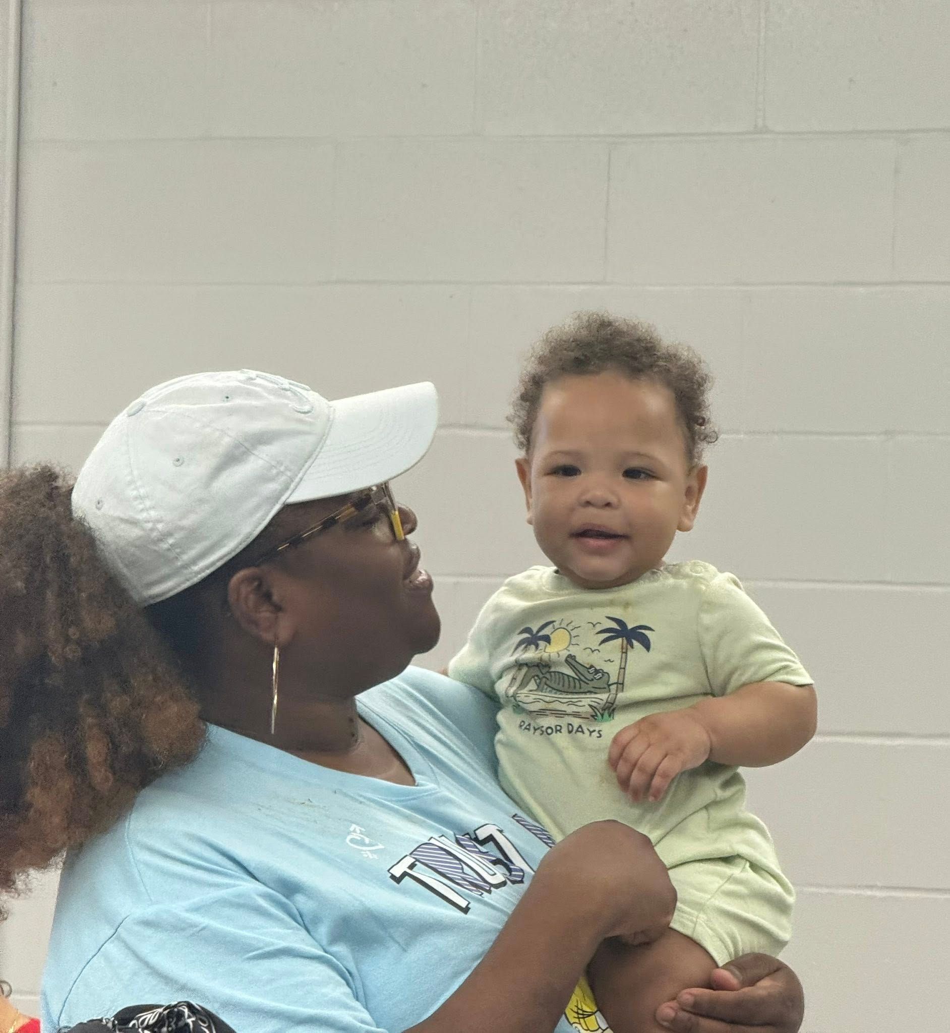 A person wearing a white baseball cap and blue shirt holds a smiling baby wearing a light green onesie against a wall.