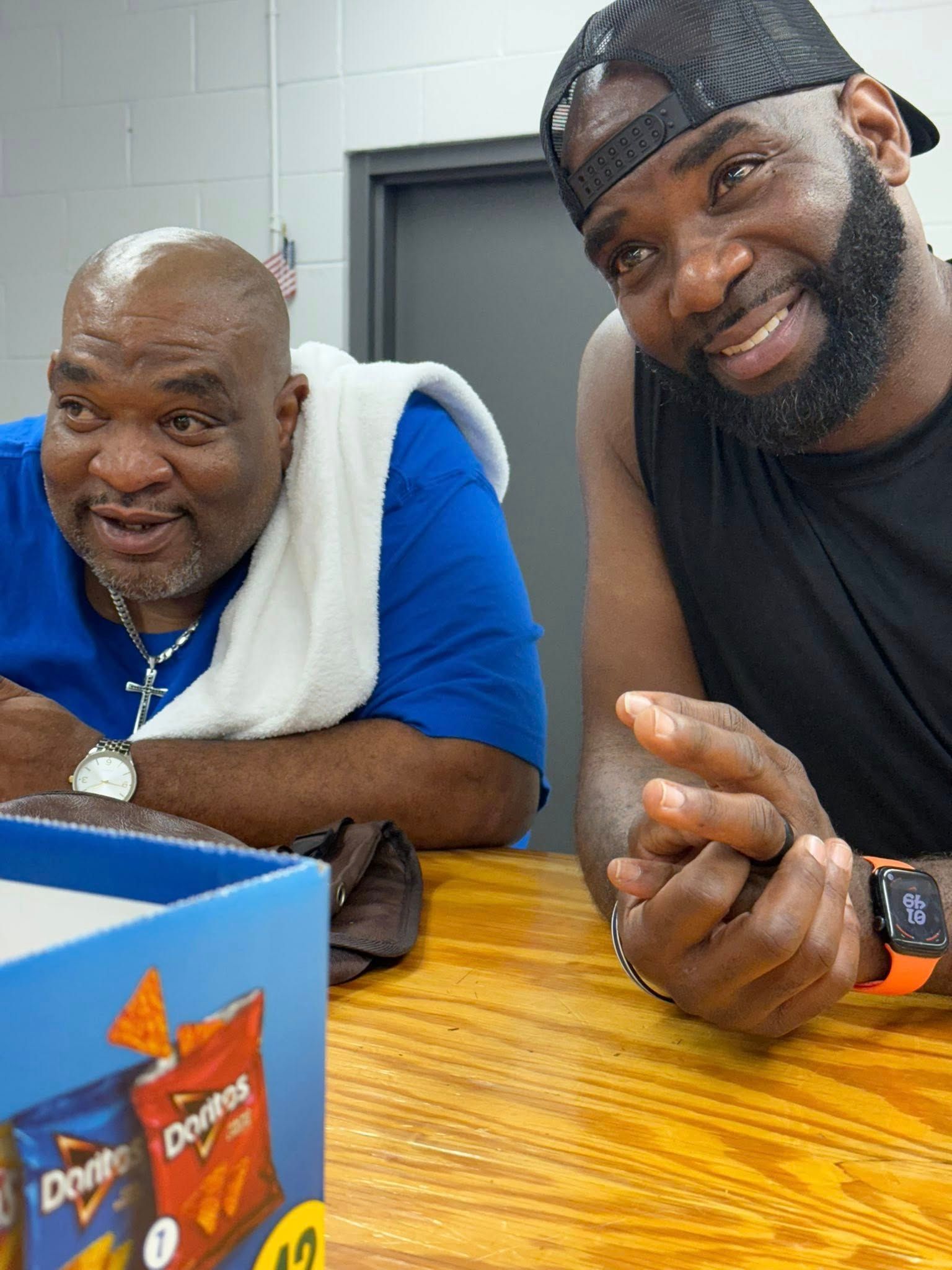 Two men sit at a table smiling, with a box of Doritos in the foreground. One wears a blue shirt and the other a tank top.