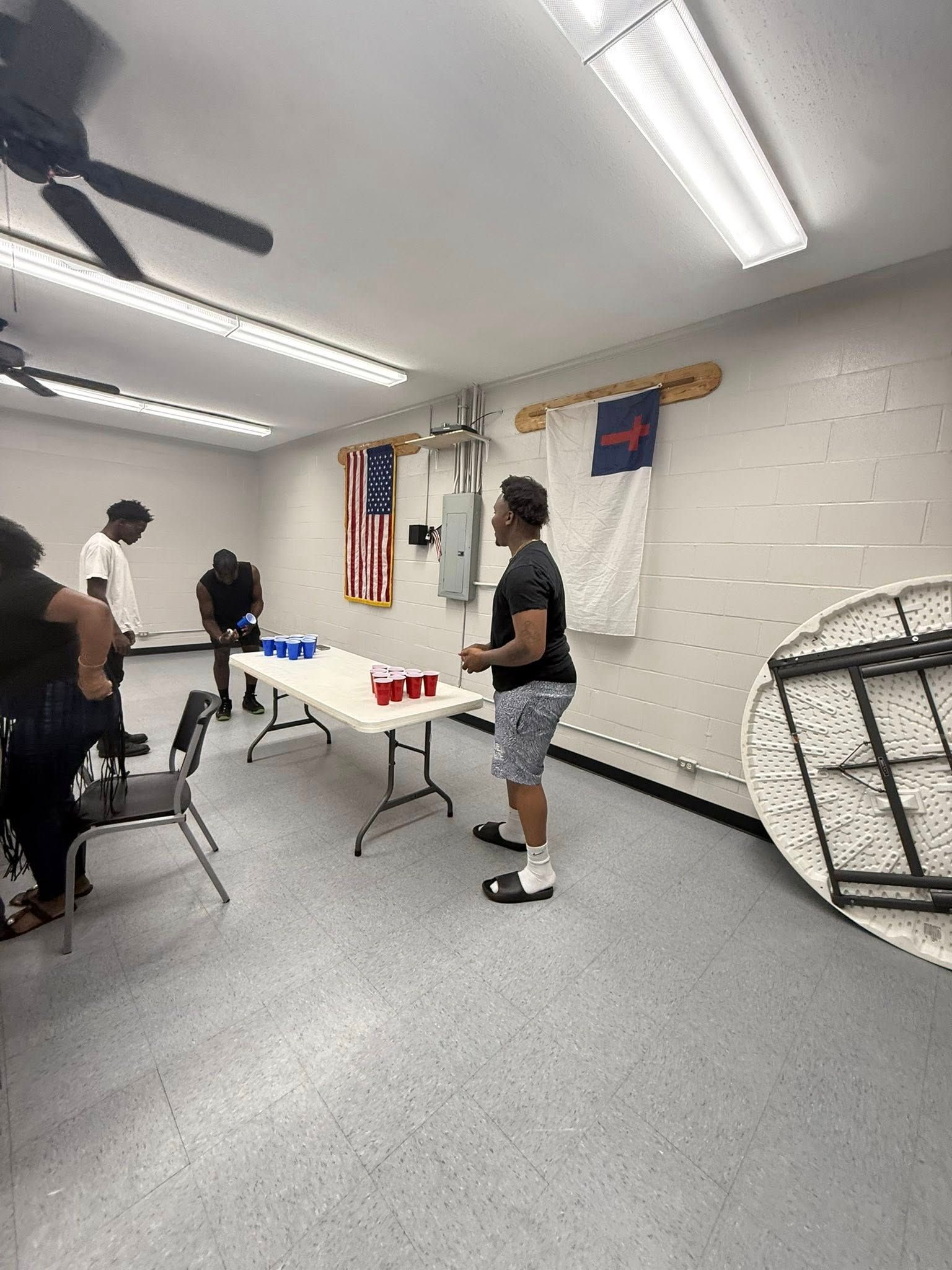 Four people in a room play beer pong at a folding table, with a flag and a folded table leaning against the wall.
