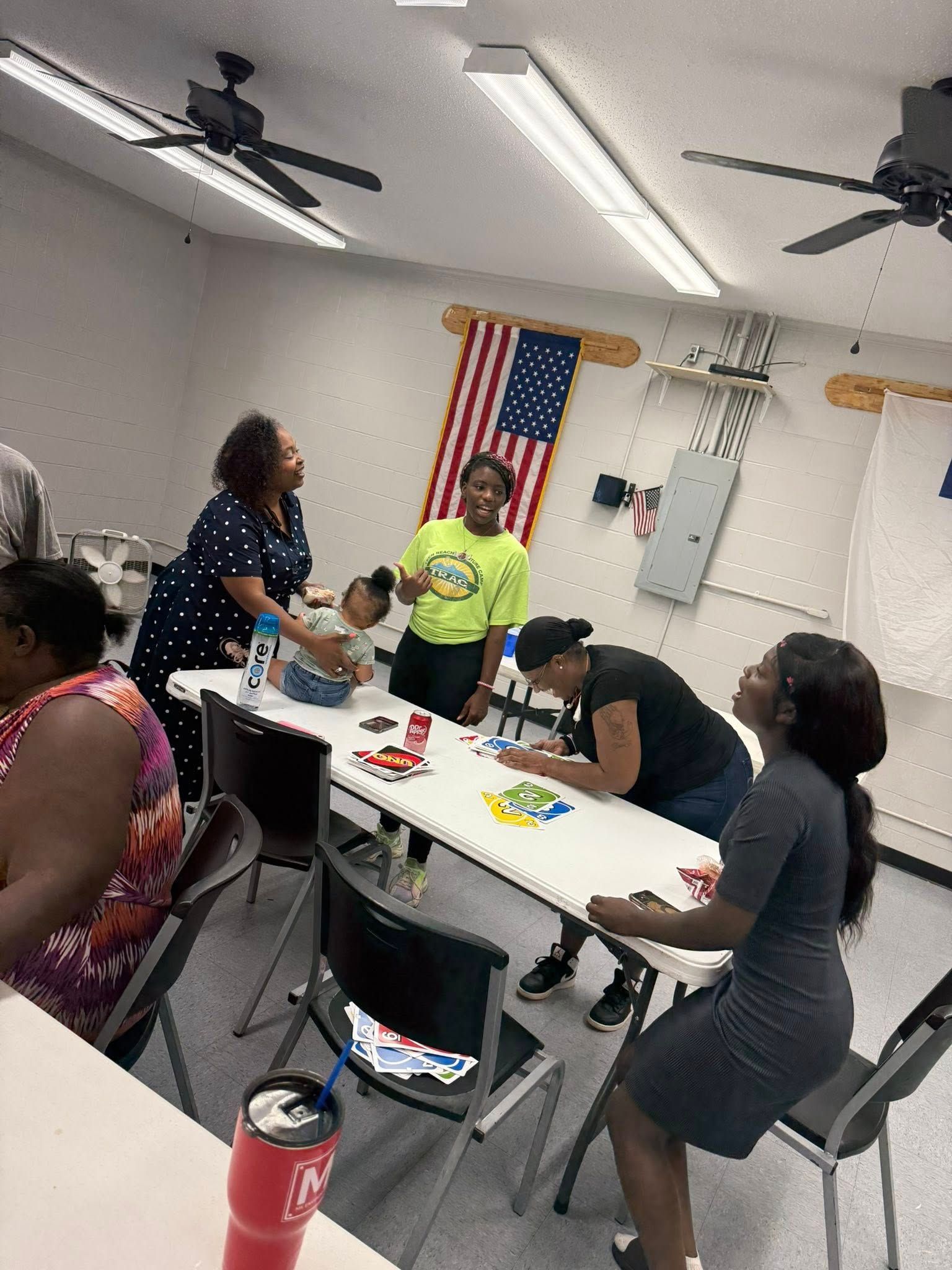 People gather around folding tables in a room with a flag on the wall, participating in a group activity or meeting.