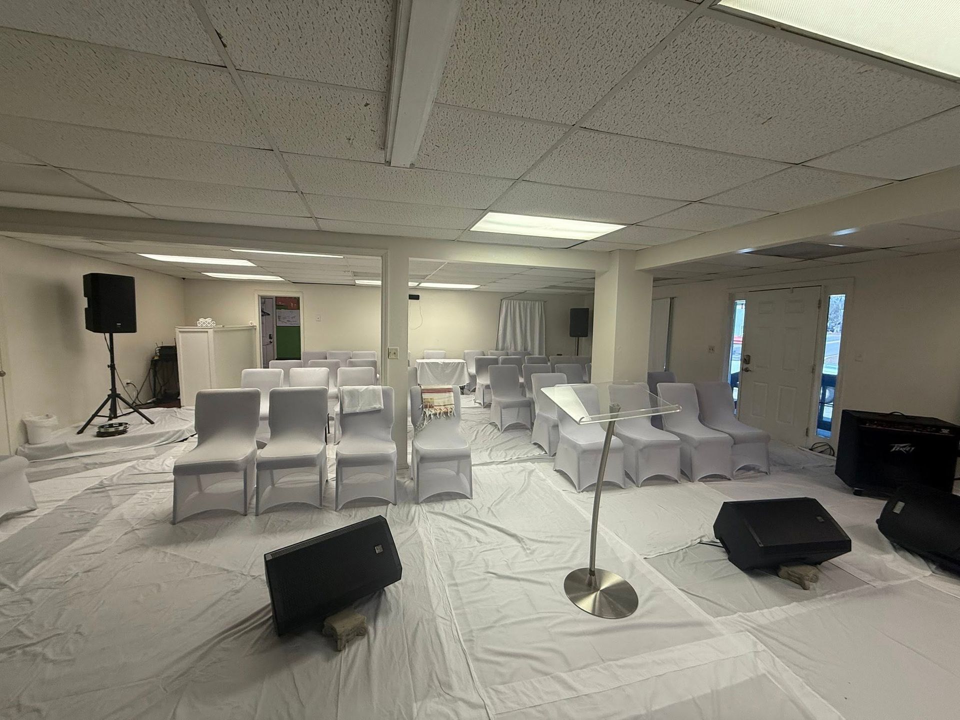 An event space set up with rows of white-covered chairs, audio equipment, and a floor covered in white fabric.