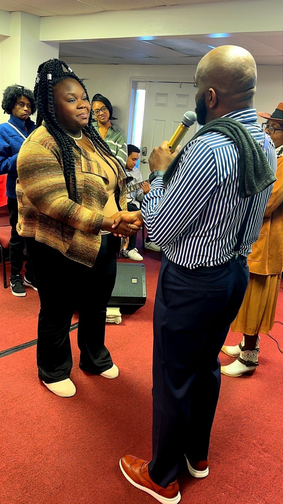 Two people shake hands at an indoor gathering; one holds a microphone while facing the other person.