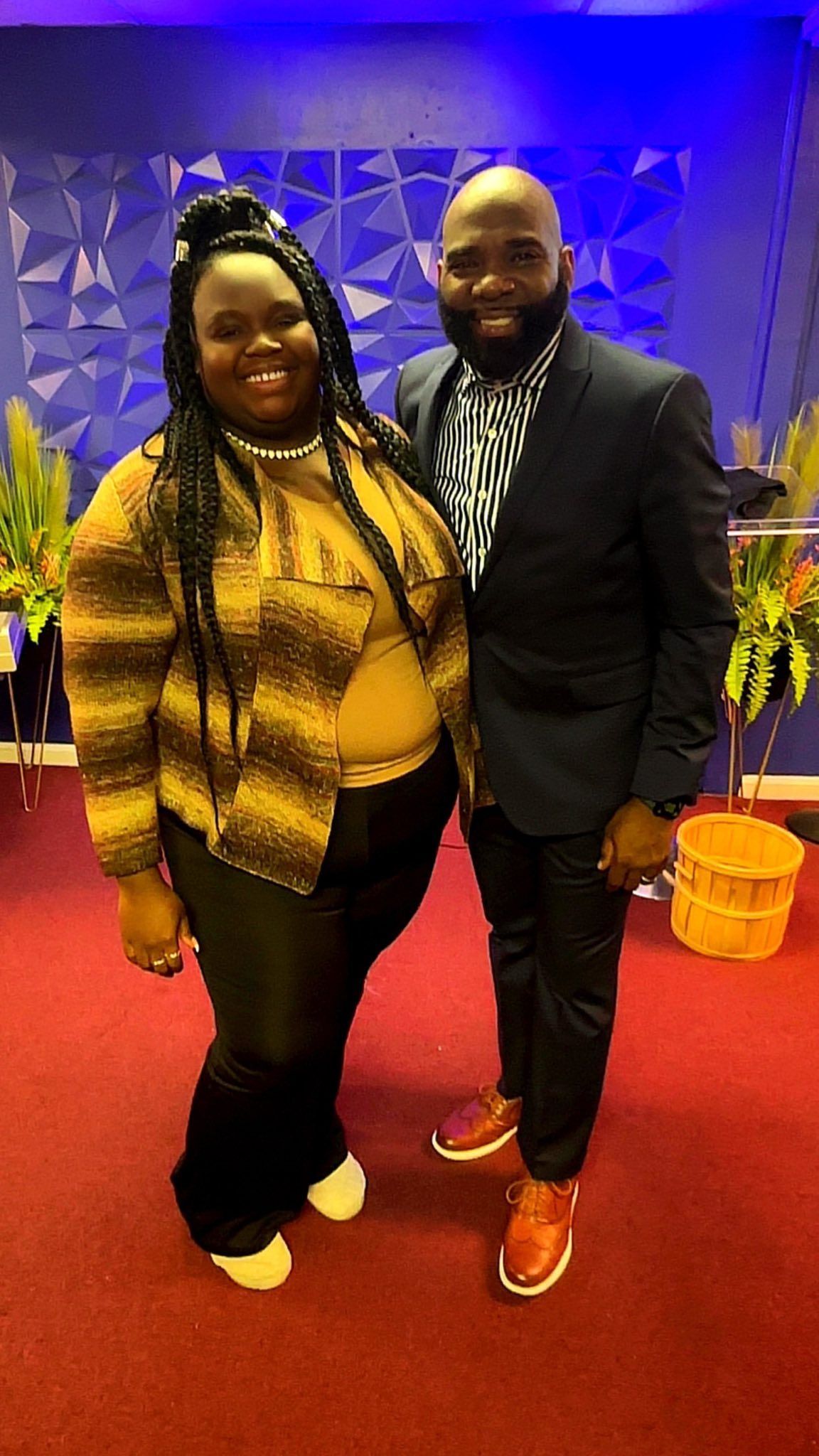 A woman and a man standing together, smiling, on a red carpet in front of a blue wall and potted plants.