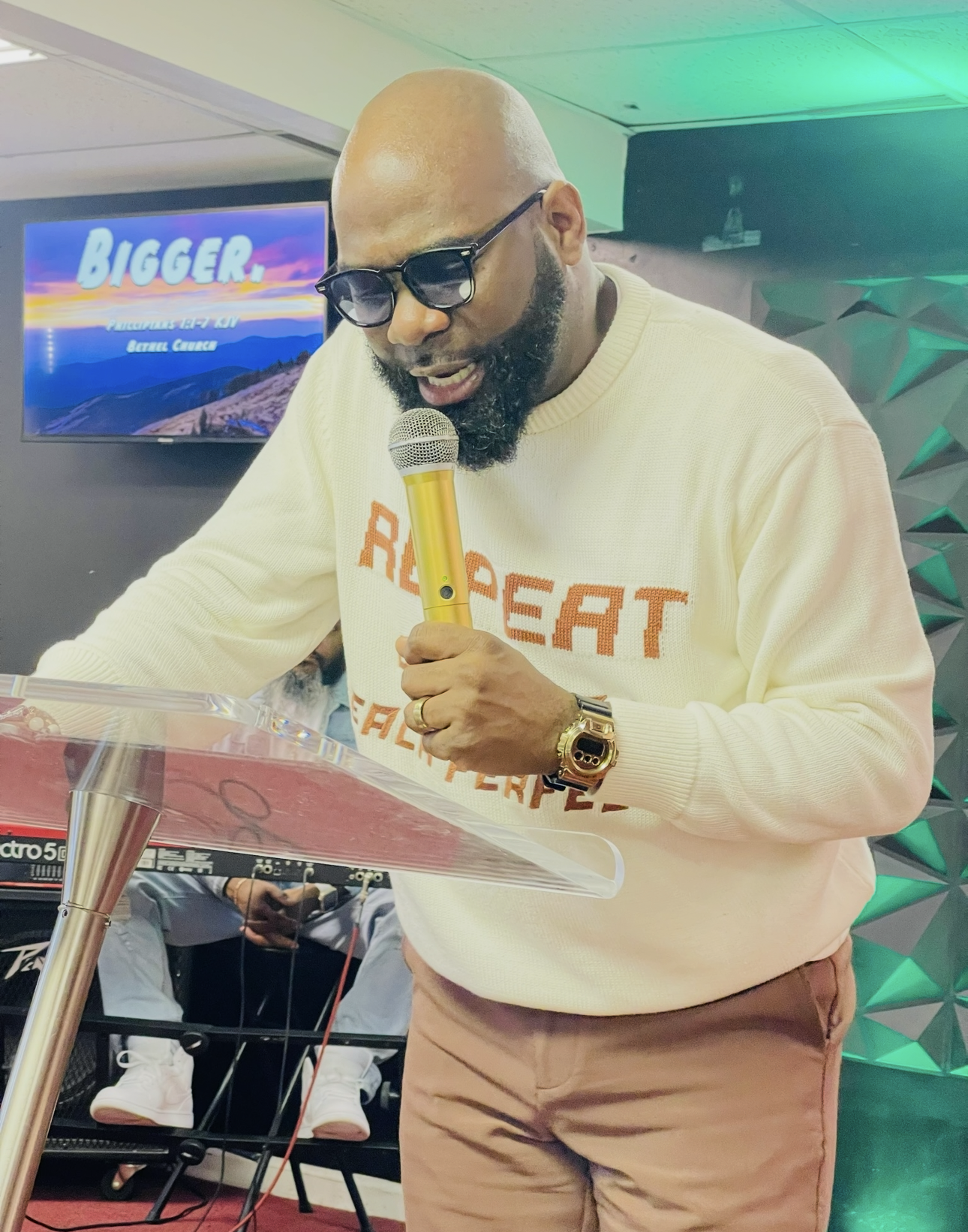 A person wearing a cream sweater speaks into a gold microphone while standing behind a podium in a room.