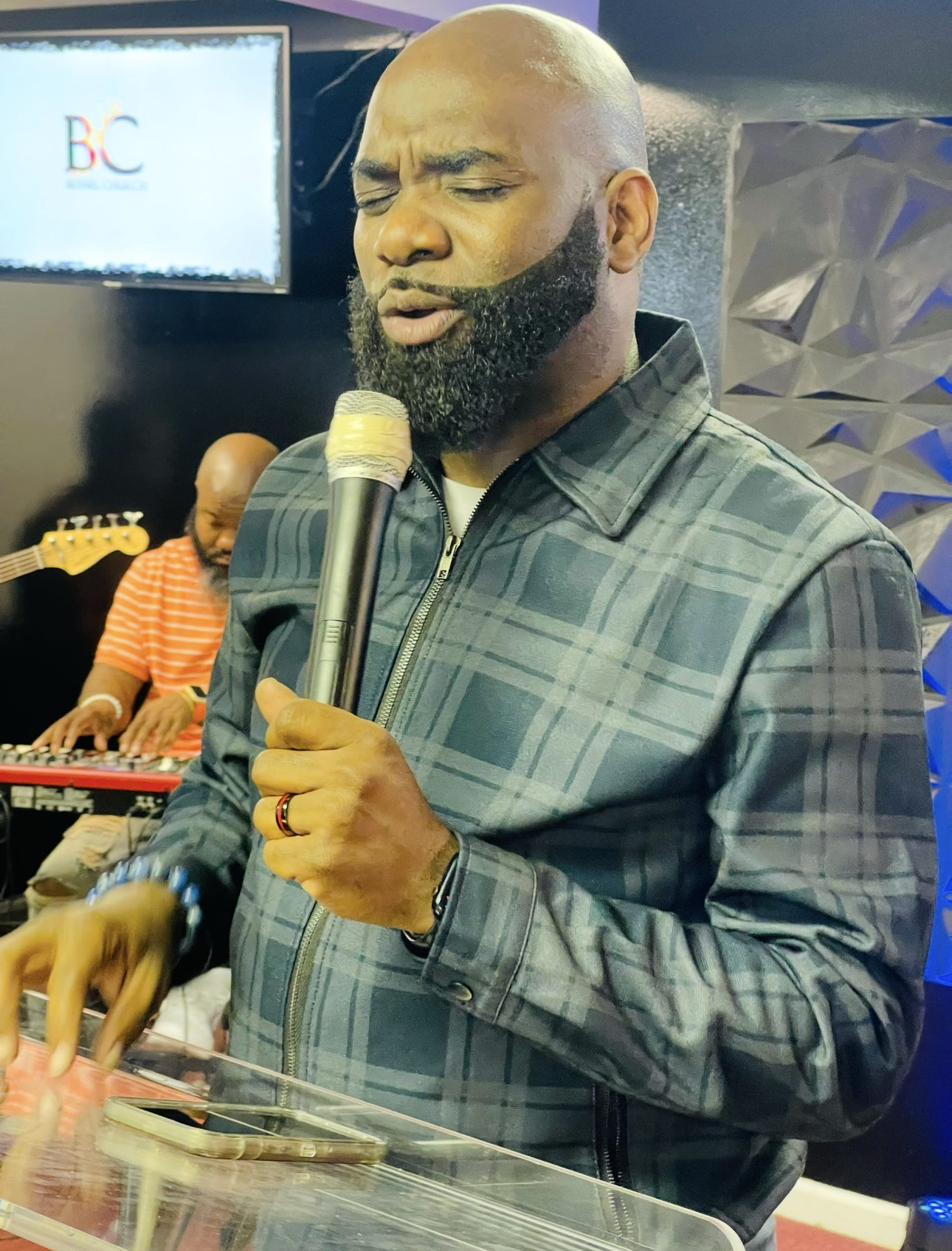 A person with a beard speaks into a microphone behind a transparent podium, with a musician playing a keyboard nearby.