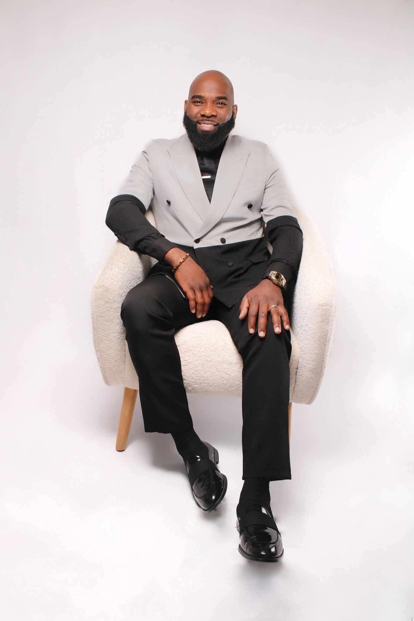 A smiling person with a beard sits in a white armchair, wearing a grey short-sleeve blazer over a black top and trousers.
